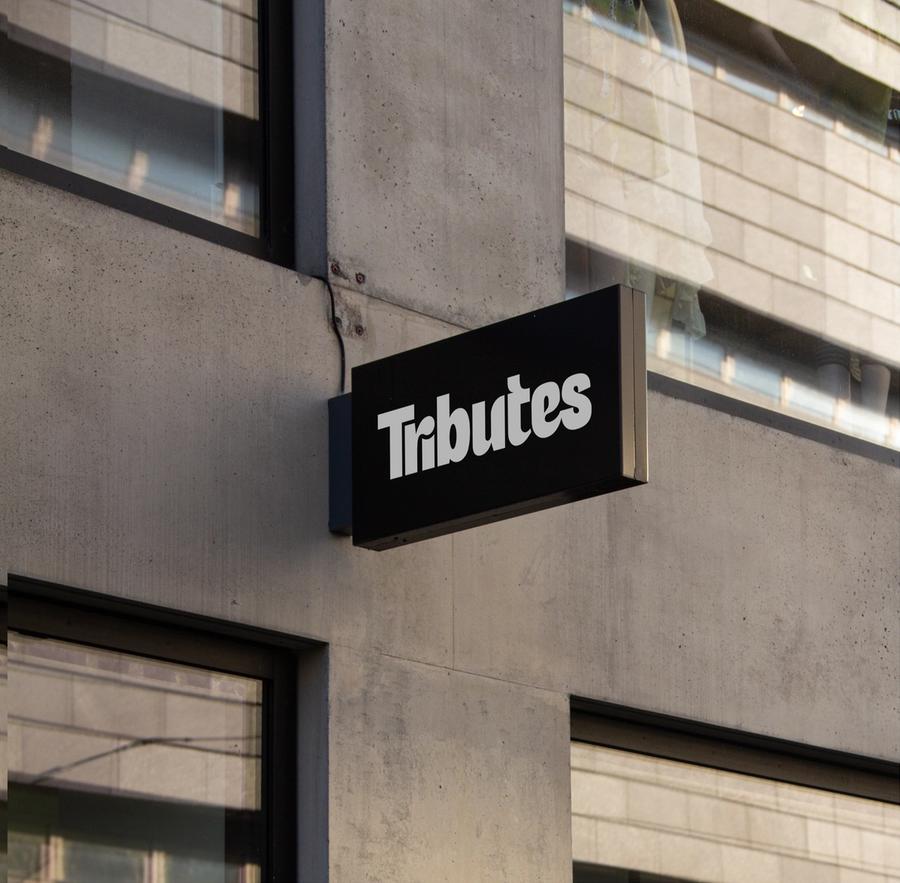 A black blade sign with the white Tributes wordmark mounted on a concrete building.