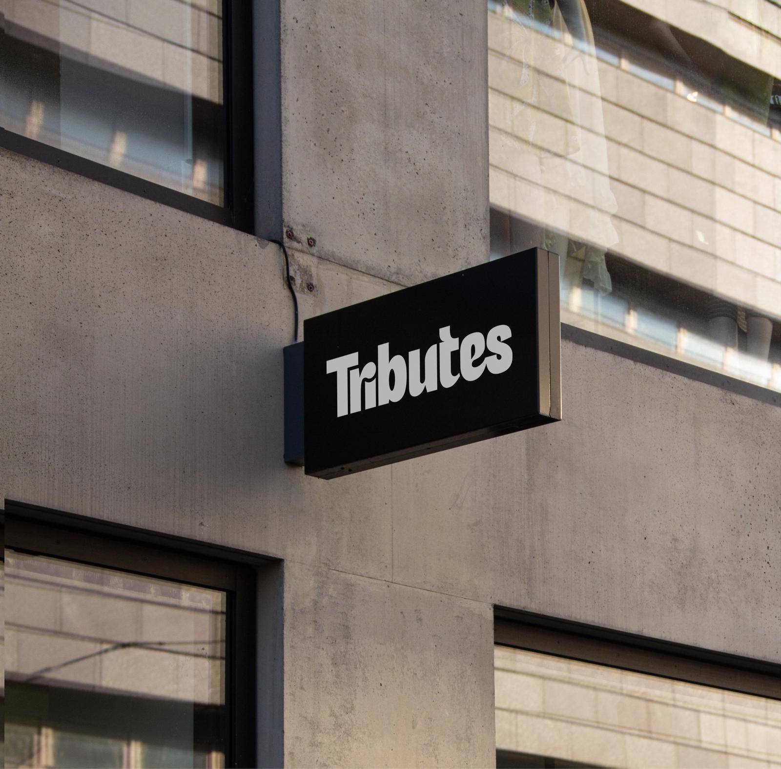 A black blade sign with the white Tributes wordmark mounted on a concrete building.