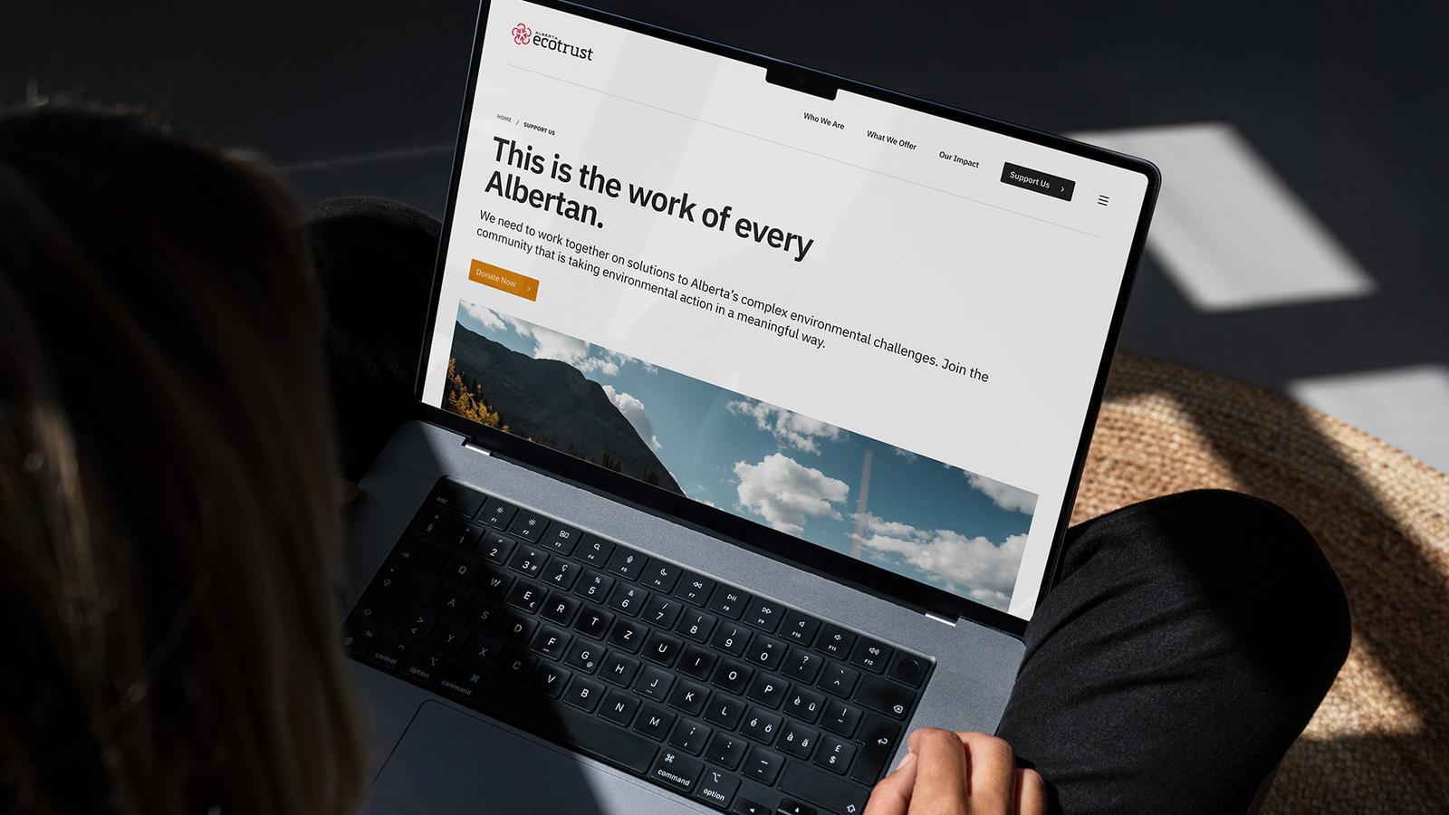 MacBook on a lap displaying the Alberta Ecotrust site headlined 'This is the work of every Albertan,' with a mountain hero image and 'Donate Now' button.