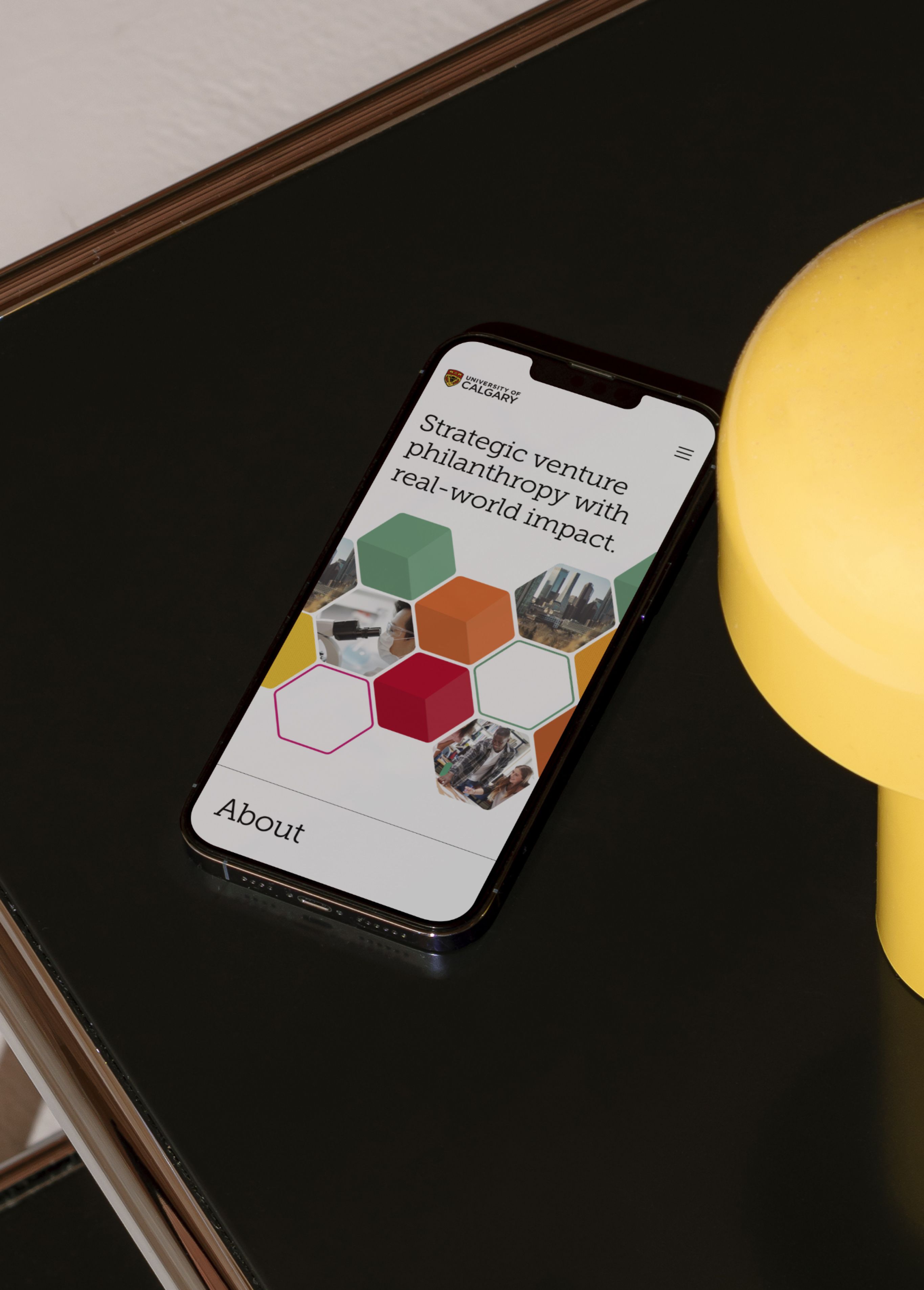 iPhone on a dark table beside a yellow lamp, showing a University of Calgary page headlined 'Strategic venture philanthropy with real-world impact' with a hexagon photo collage.
