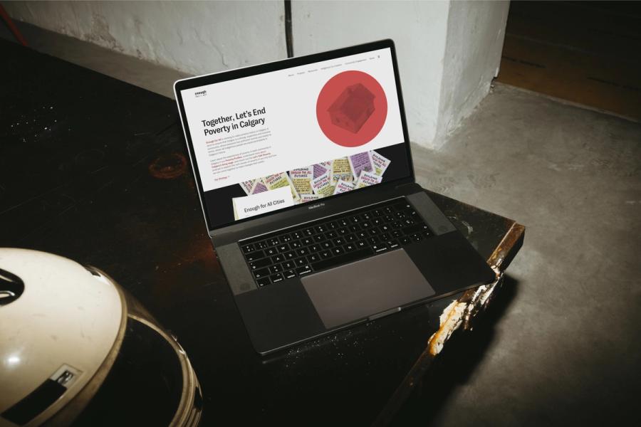 MacBook Pro on a dark worn surface showing the Enough for All website headlined 'Together, Let's End Poverty in Calgary,' with a red house illustration.