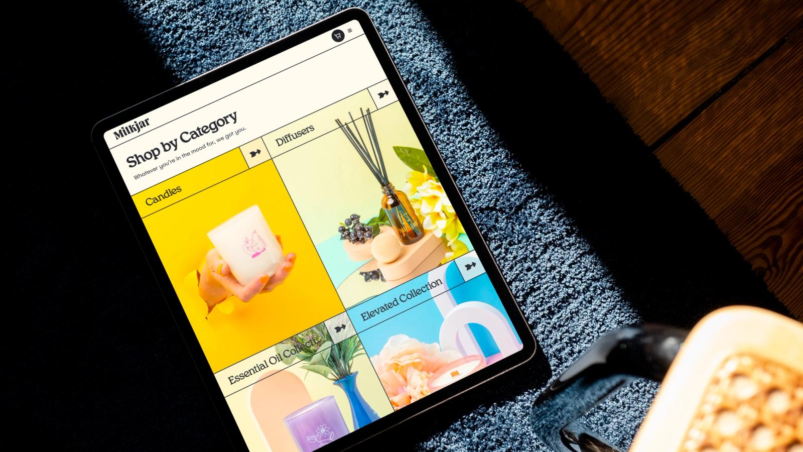 Tablet on a blue rug displaying the Milkjar 'Shop by Category' page with colorful tiles for Candles, Diffusers, Essential Oil and Elevated collections.