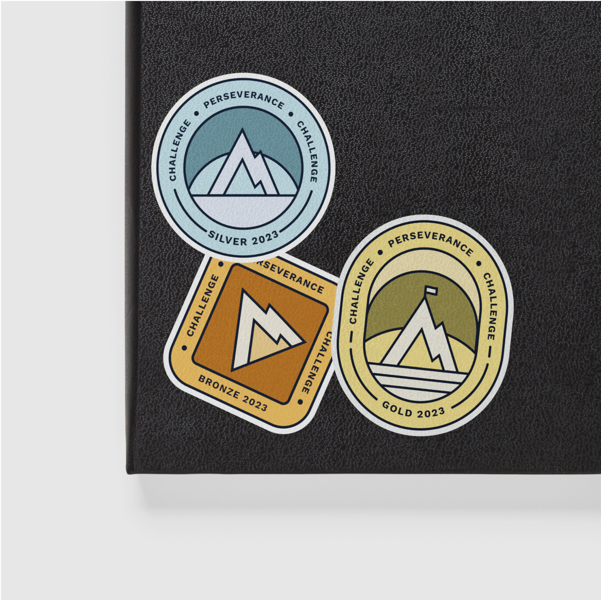 Three mountain-themed achievement badge stickers reading 'Silver 2023,' 'Bronze 2023,' and 'Gold 2023,' arranged on a black notebook cover.