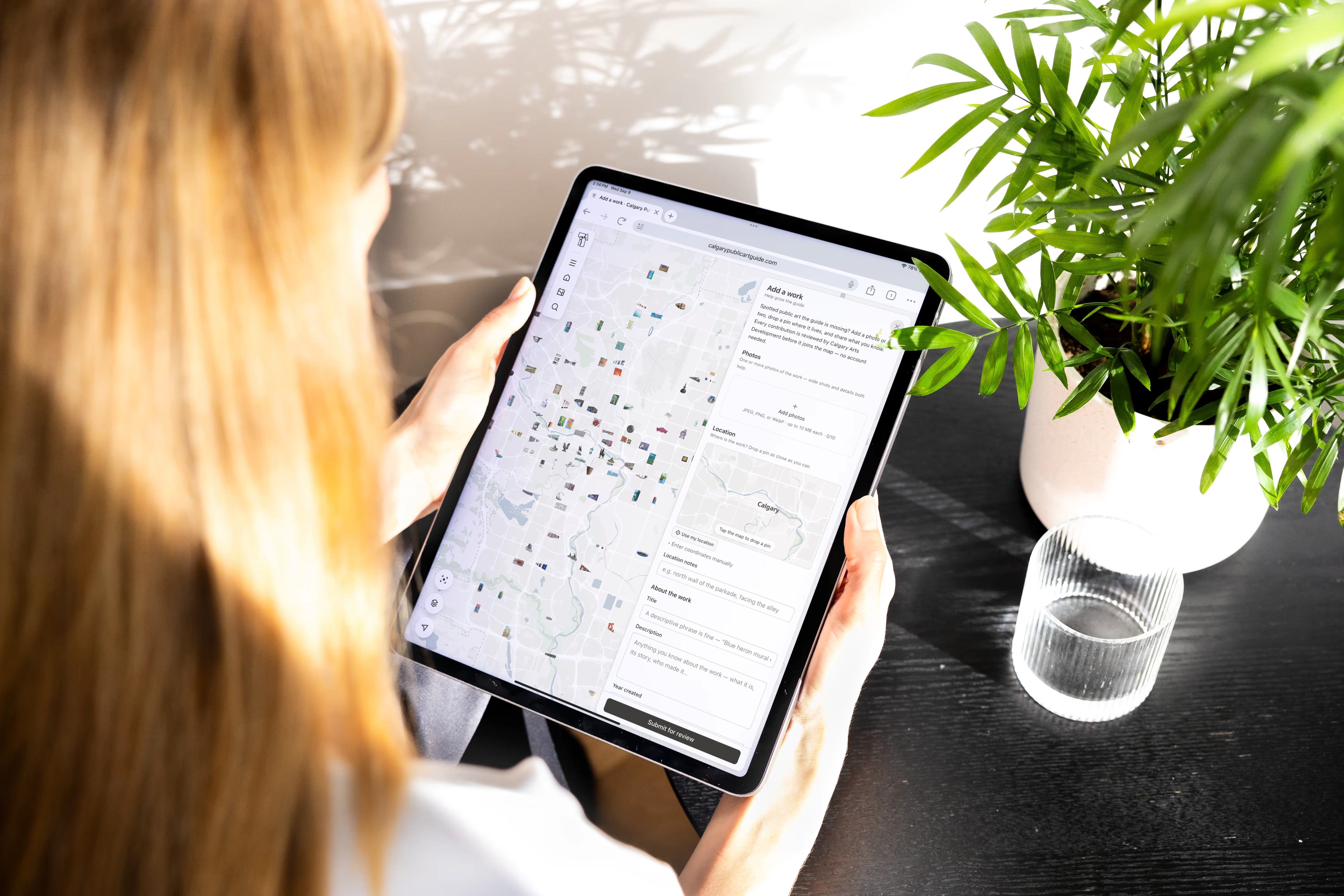 A person holding a tablet showing the Calgary Public Art Guide map beside a potted plant.