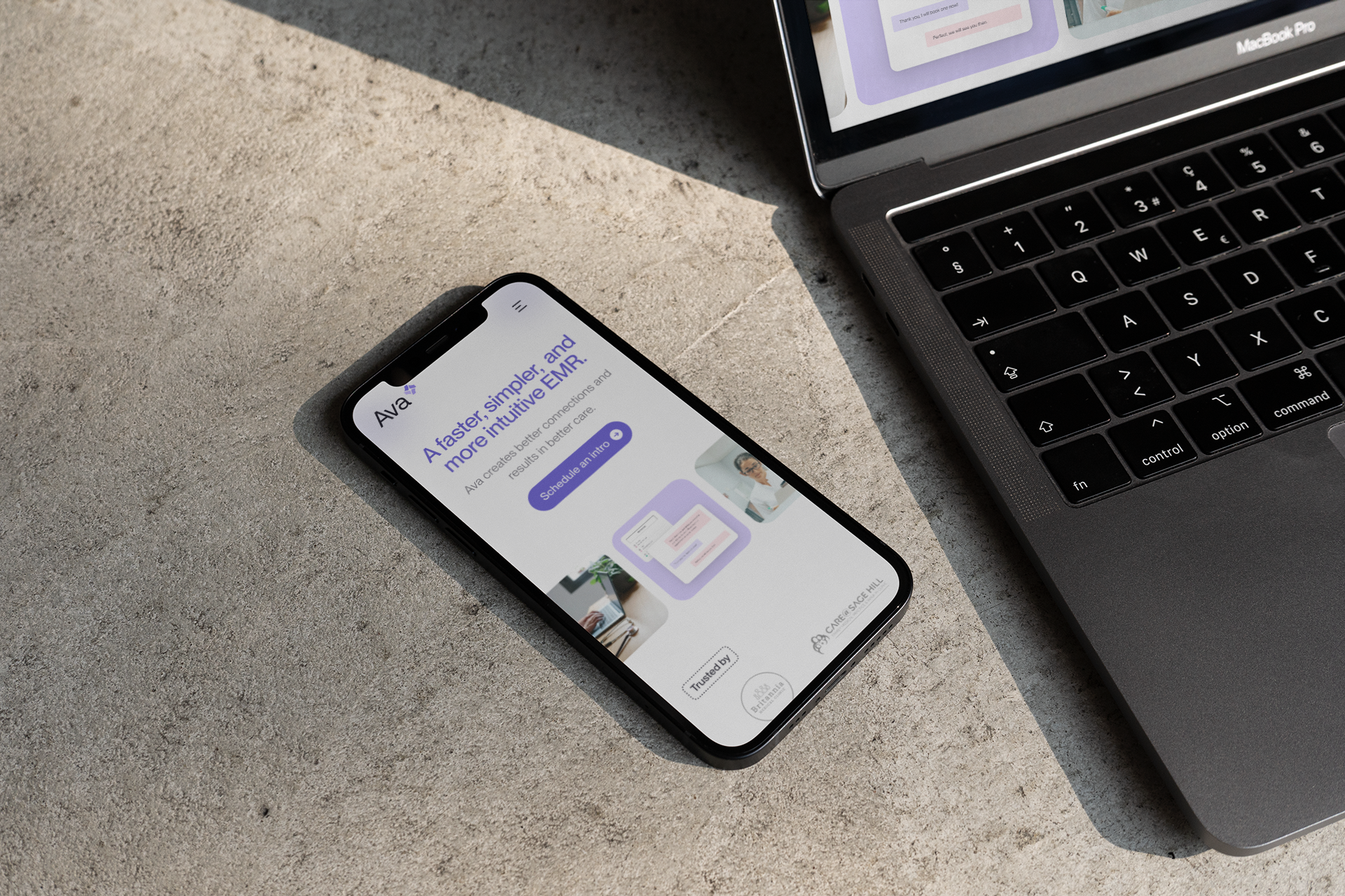 Smartphone on a concrete surface beside a MacBook Pro, the phone displaying the Ava website headlined 'A faster, simpler, and more intuitive EMR.'