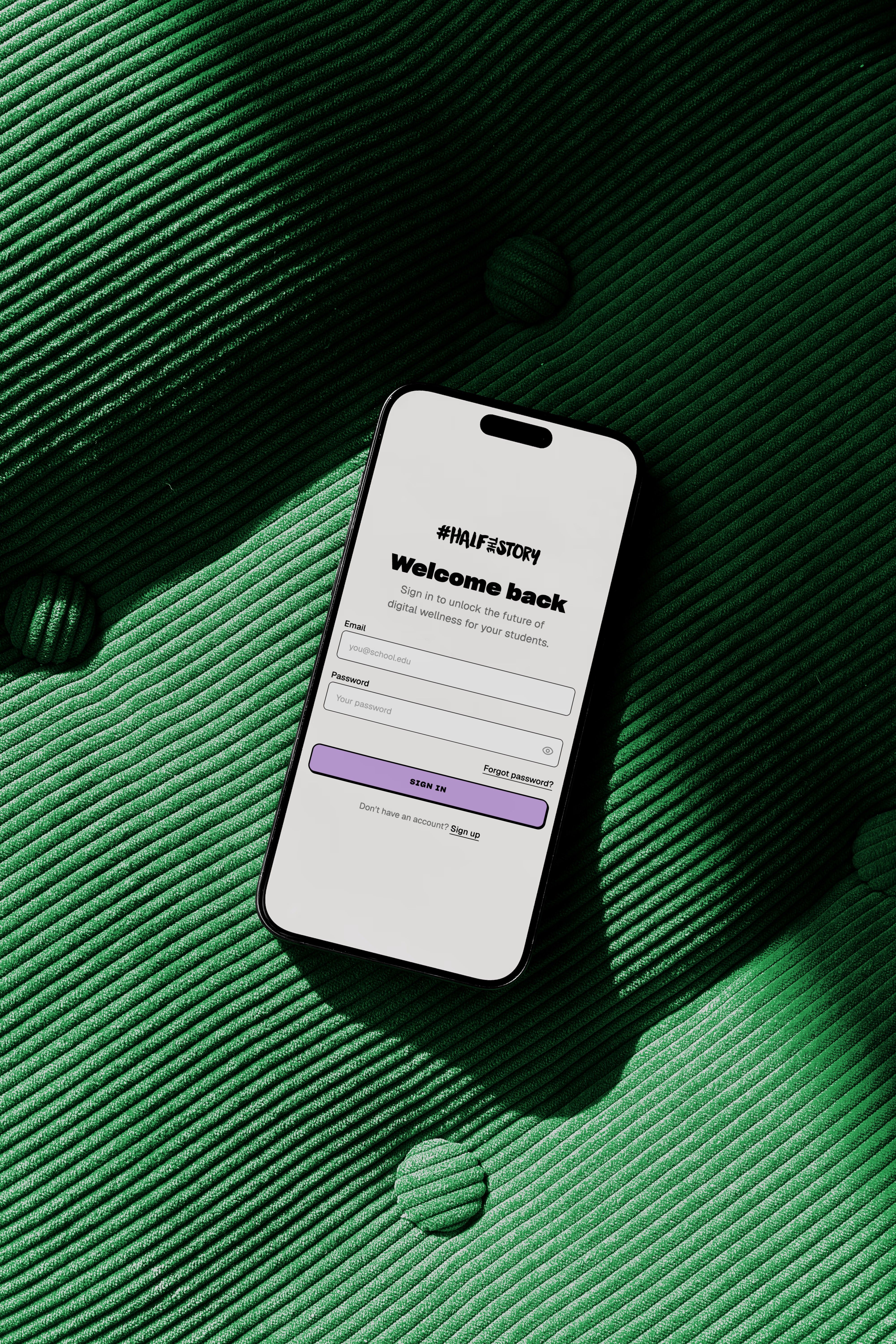 A phone lying on green corduroy, showing the #HalfTheStory learning platform sign-in screen headed Welcome back.
