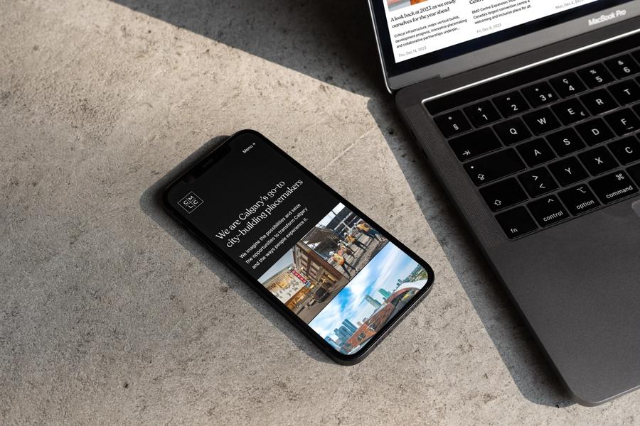 iPhone on a concrete surface beside a MacBook Pro, the phone showing a dark CMLC webpage reading 'We are Calgary's go-to city-building placemakers.'