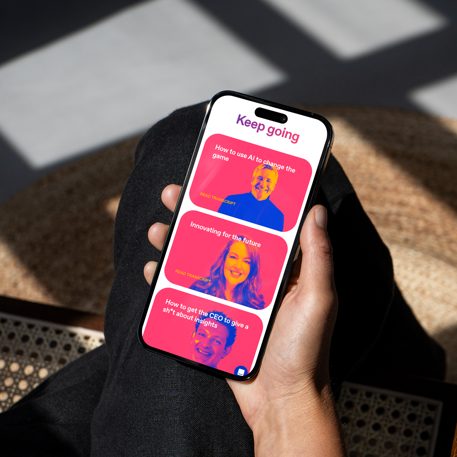 Hand holding an iPhone in sunlight, the screen showing a pink 'Keep going' content feed with duotone speaker portraits and article titles.