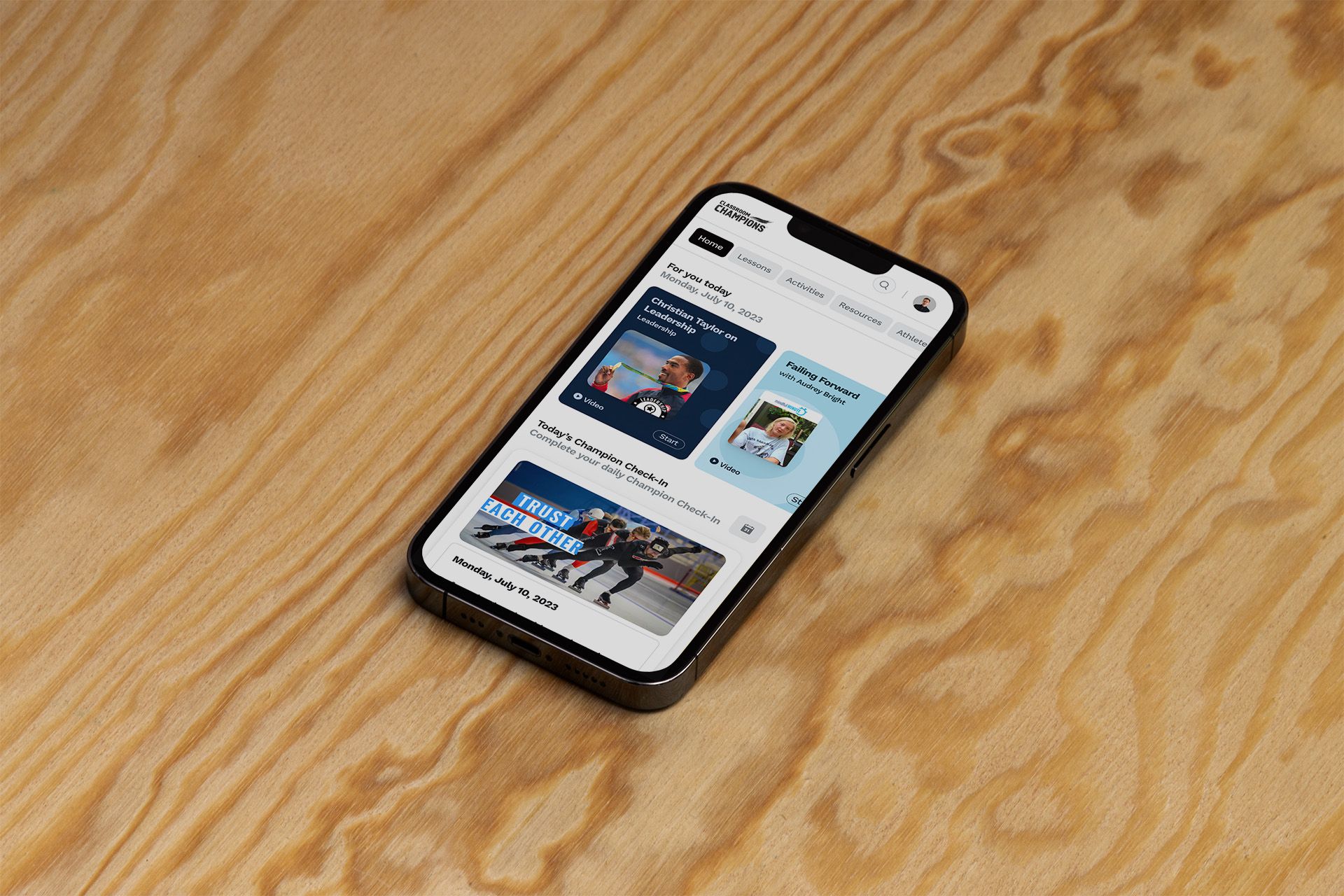 iPhone on a wood surface showing the Classroom Champions app home screen with 'For you today' video lessons and a 'Champion Check-In' card.