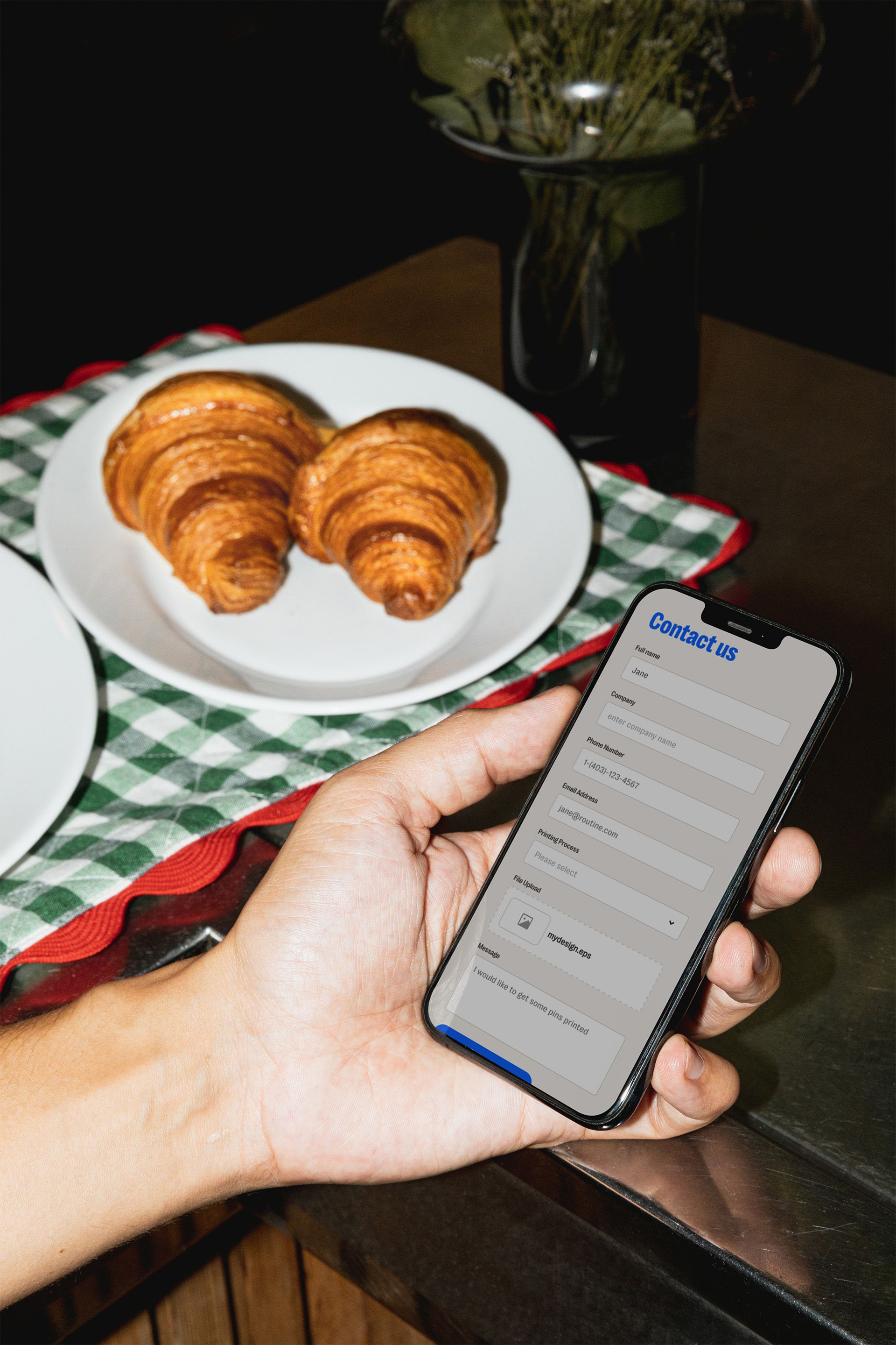 A hand holding a phone showing the Tributes contact form beside a plate of croissants on a gingham napkin.