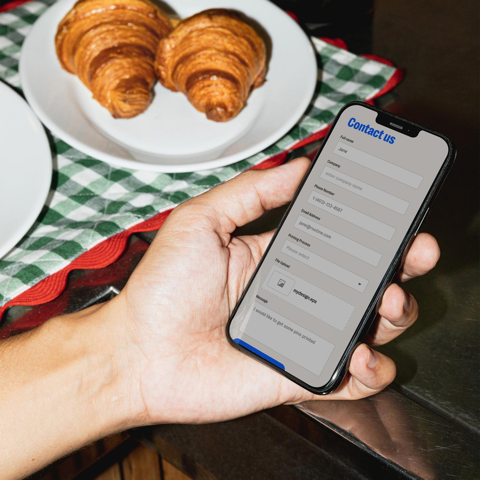 A hand holding a phone showing the Tributes contact form beside a plate of croissants on a gingham napkin.