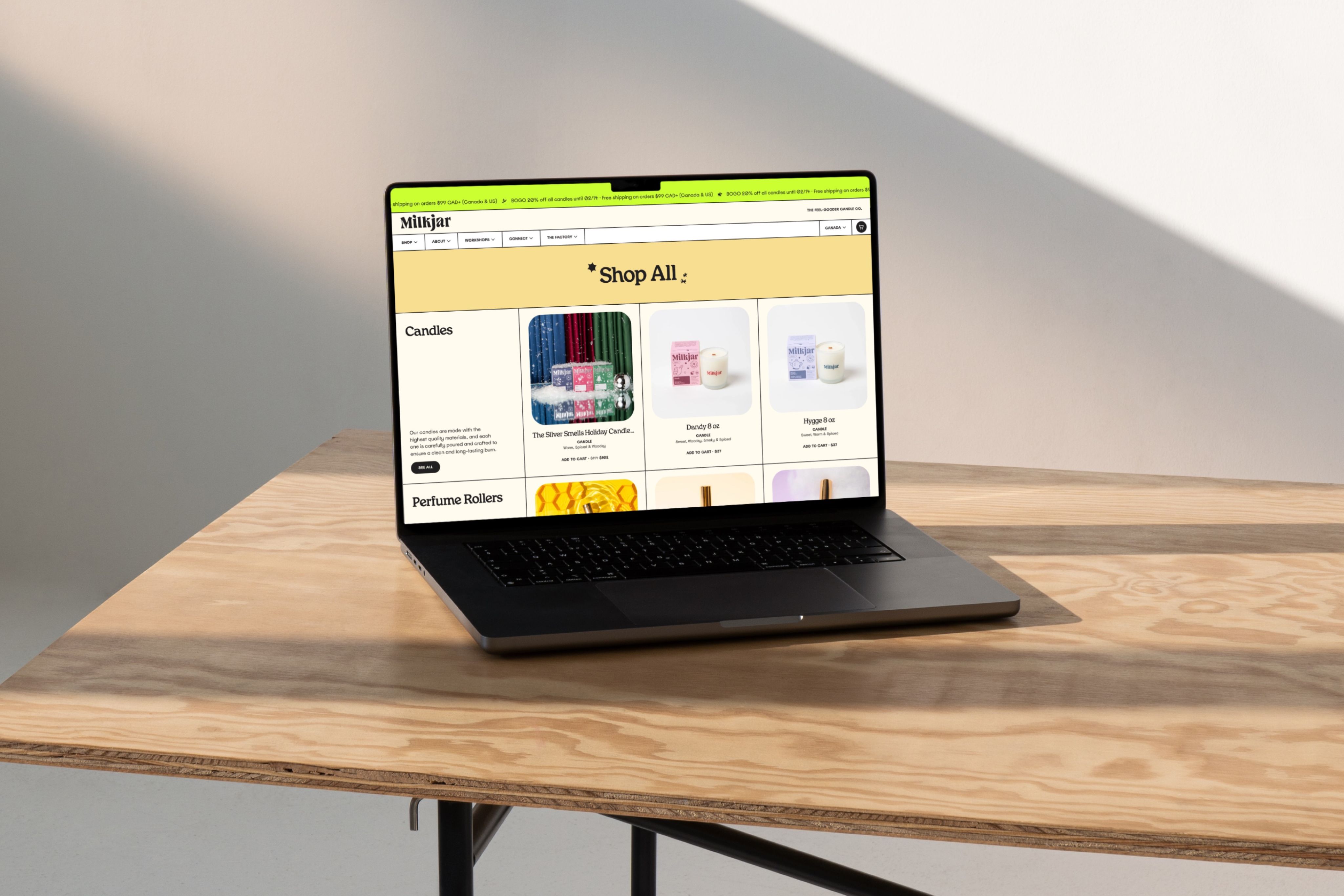 Open laptop on a light wood table showing the Milkjar online store 'Shop All' page with candle and perfume roller product listings.