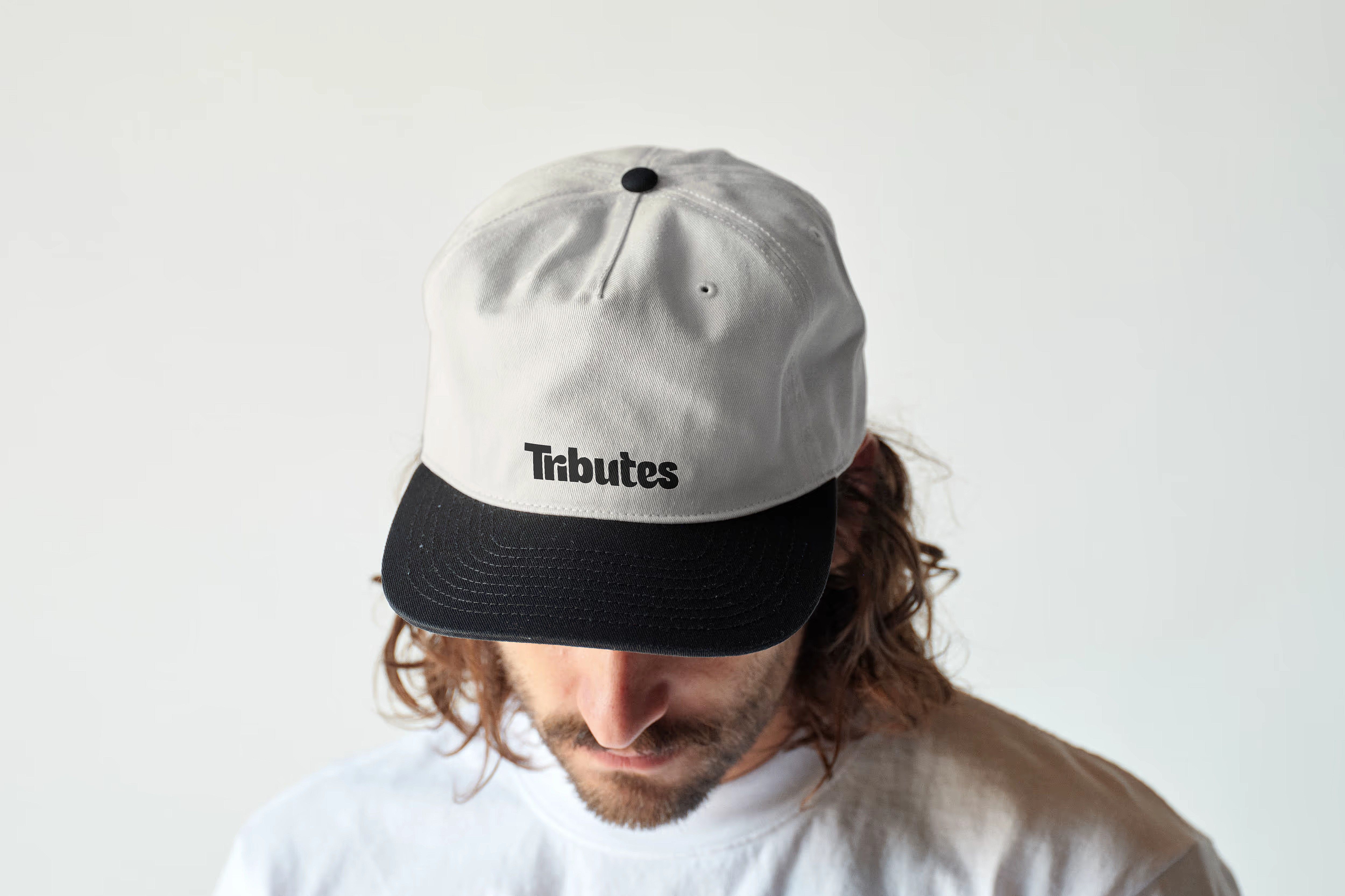 A person looking down in a cream and black cap embroidered with the Tributes wordmark.