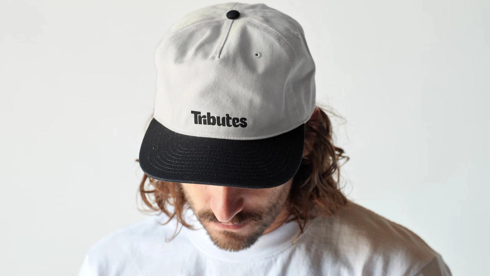 A person looking down in a cream and black cap embroidered with the Tributes wordmark.