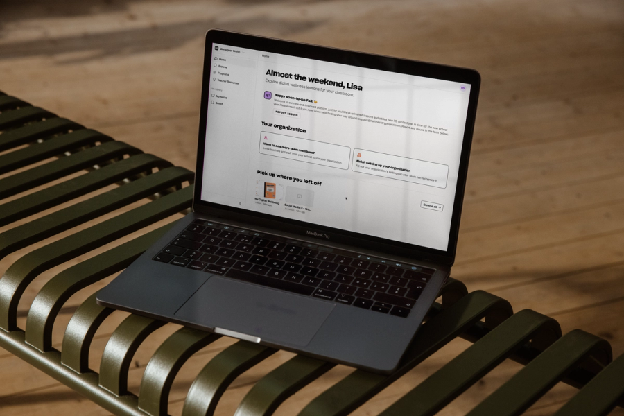 A laptop on a slatted green bench showing the Classroom Champions teacher dashboard, greeting Lisa with an Almost the weekend message.