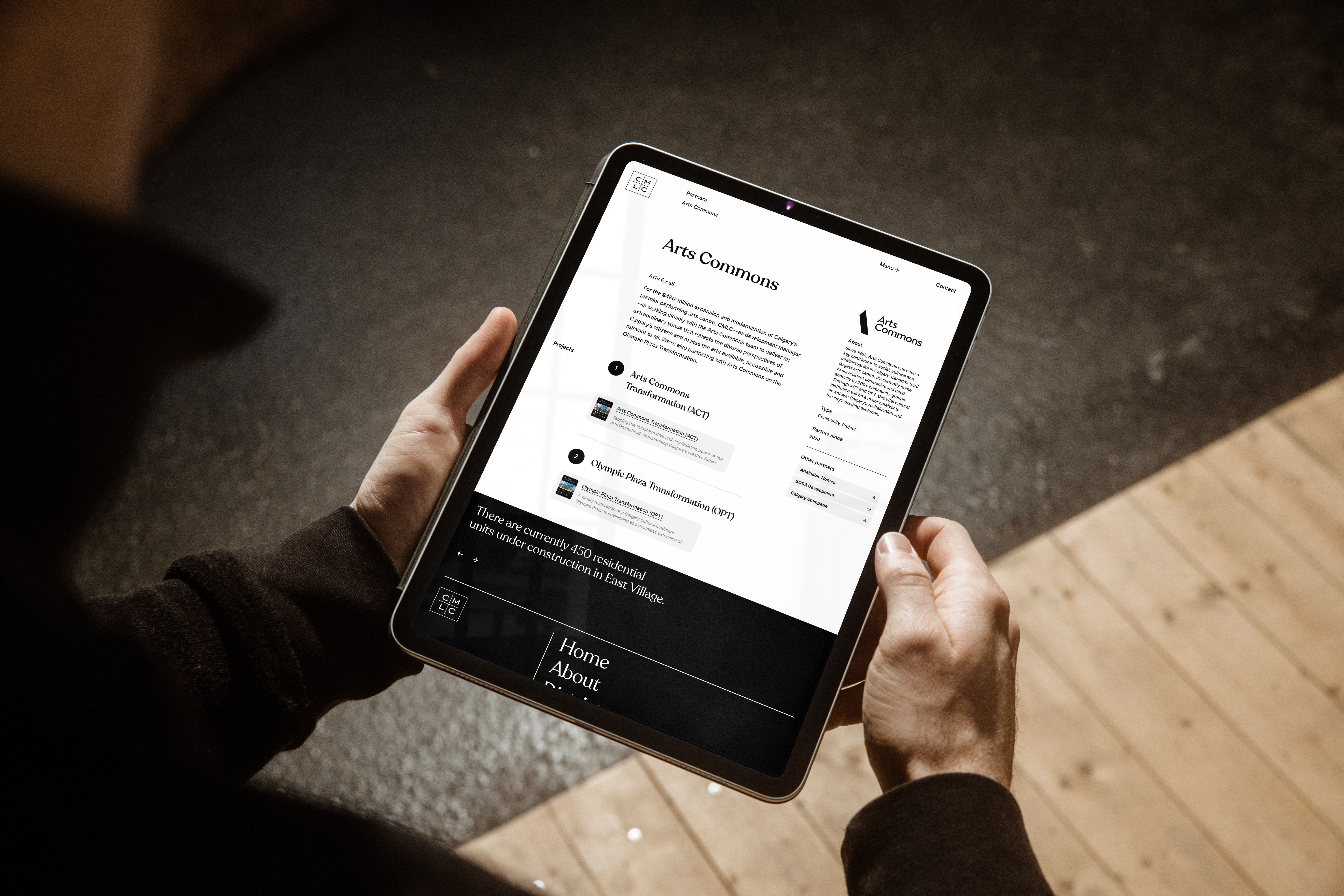 Hands holding a tablet that displays the CMLC 'Arts Commons' partner page, viewed over the shoulder against a wooden floor.