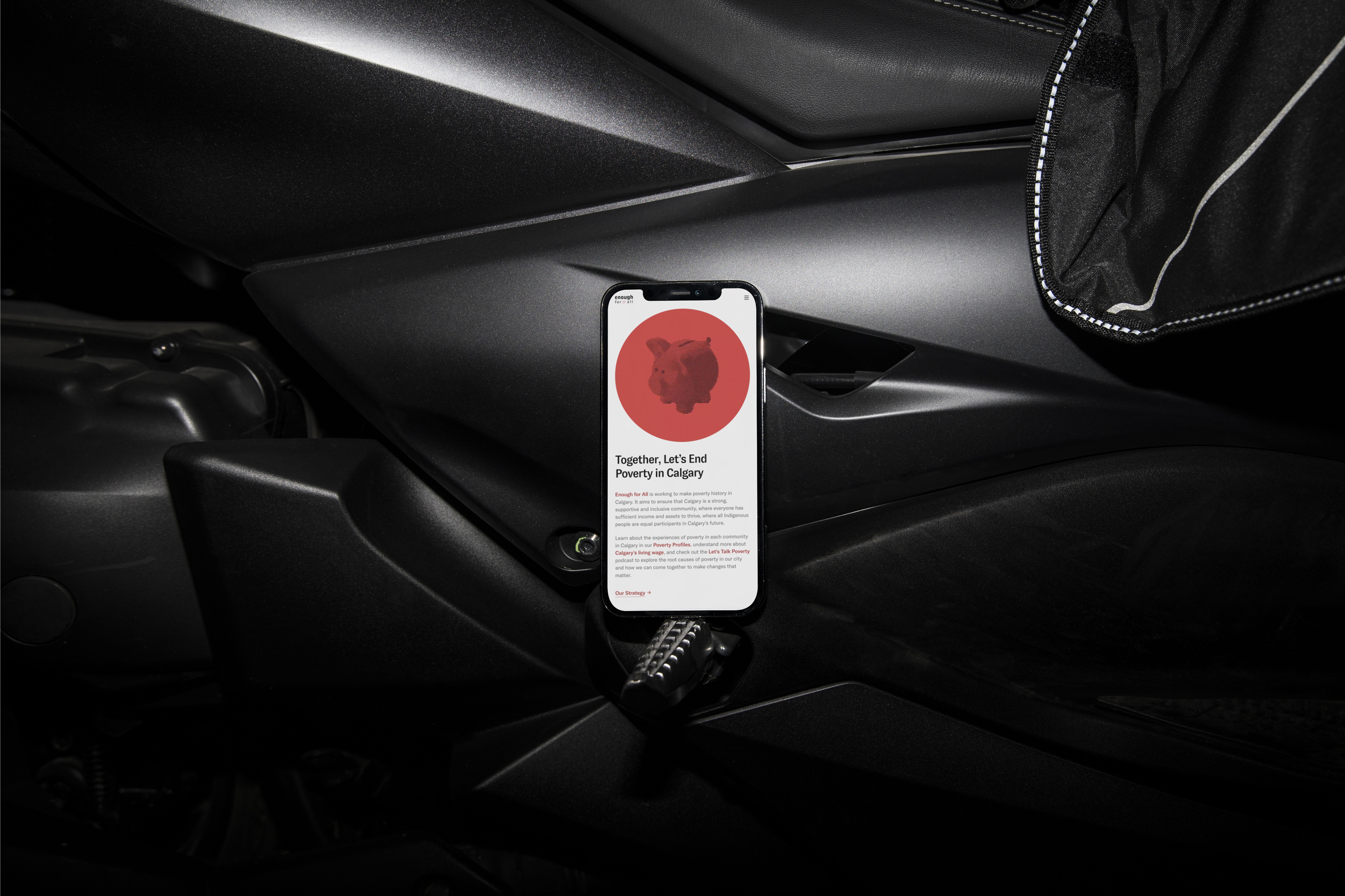 Smartphone propped in a dim car interior displaying a web page headed 'Together, Let's End Poverty in Calgary' with a red circle and piggy bank graphic.
