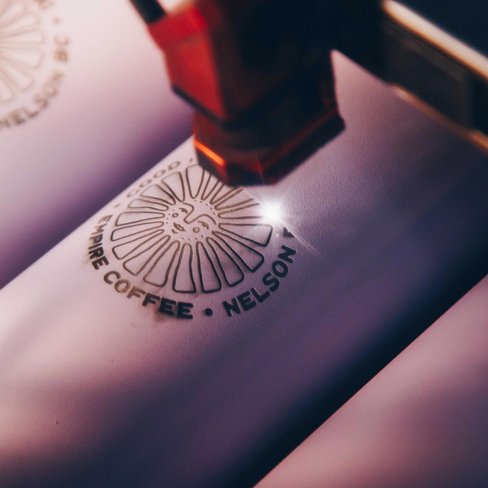 A laser engraver etching a coffee roaster's round logo onto a pink tumbler.
