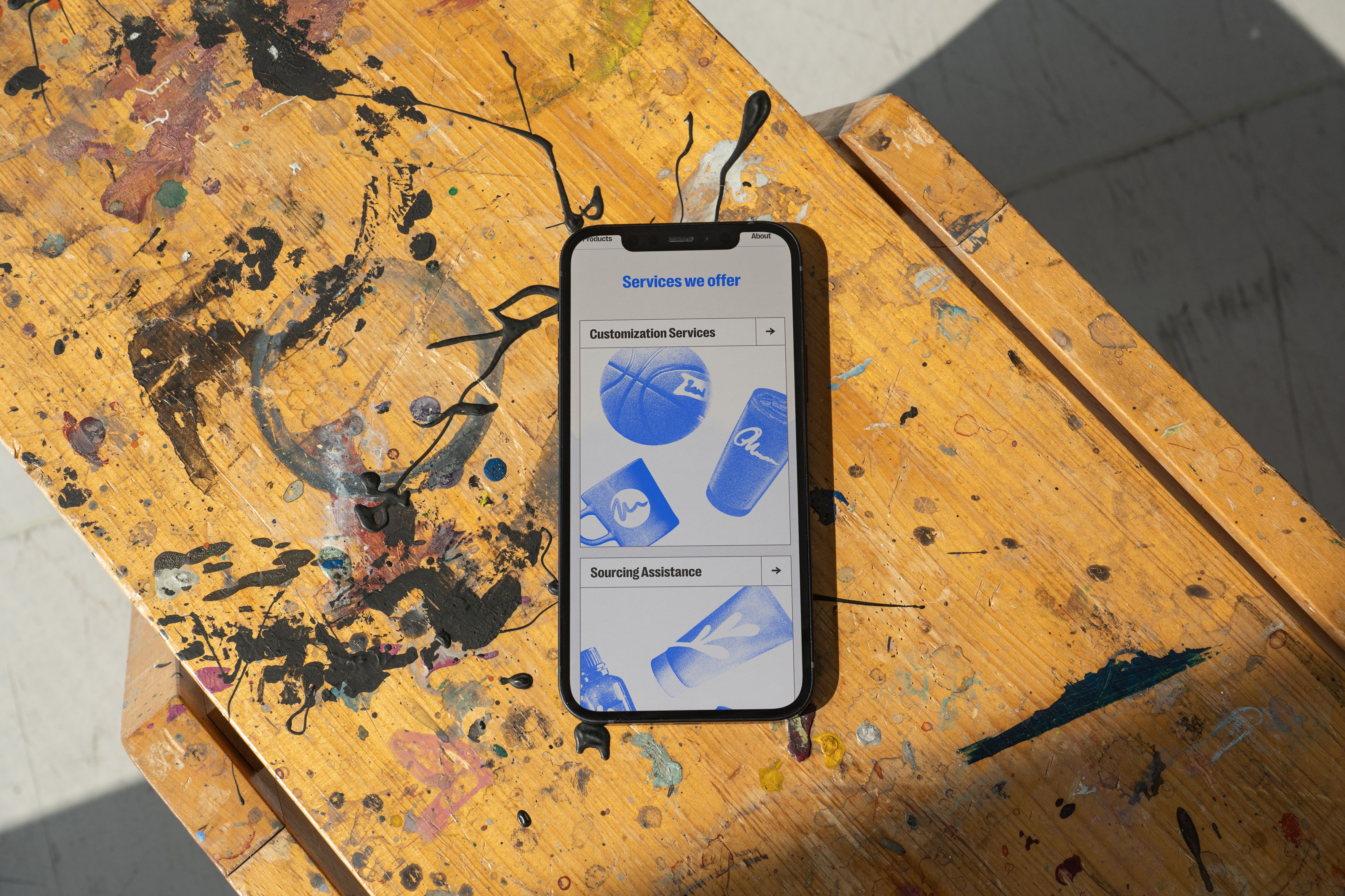 A phone on a paint-splattered wooden stool, showing the Tributes services page with customization and sourcing sections.