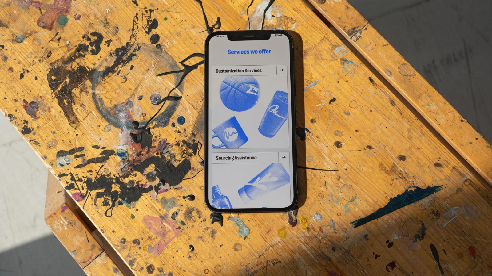 A phone on a paint-splattered wooden stool, showing the Tributes services page with customization and sourcing sections.