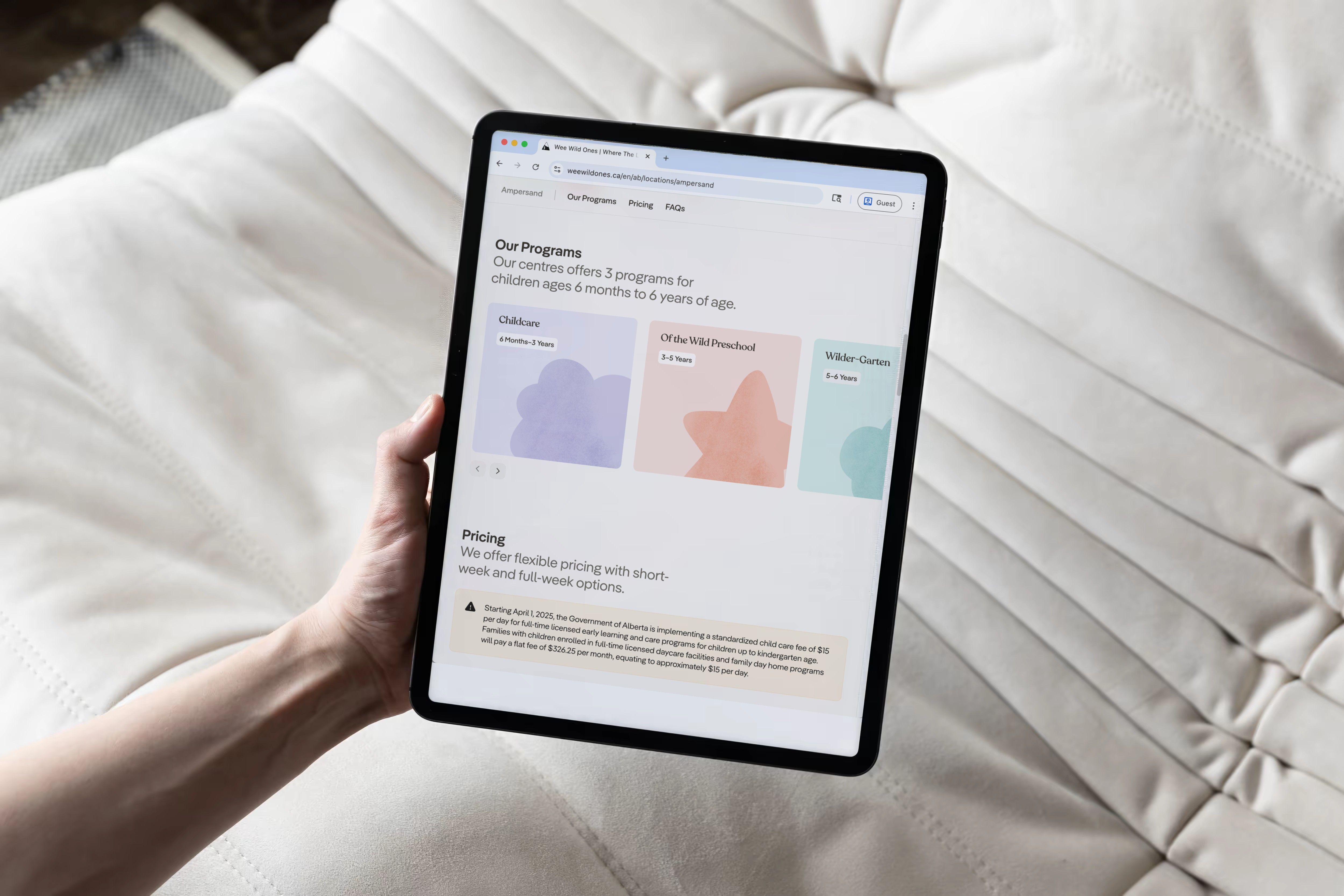 A hand holding a tablet showing the Wee Wild Ones programs and pricing sections, over a white quilted cushion.