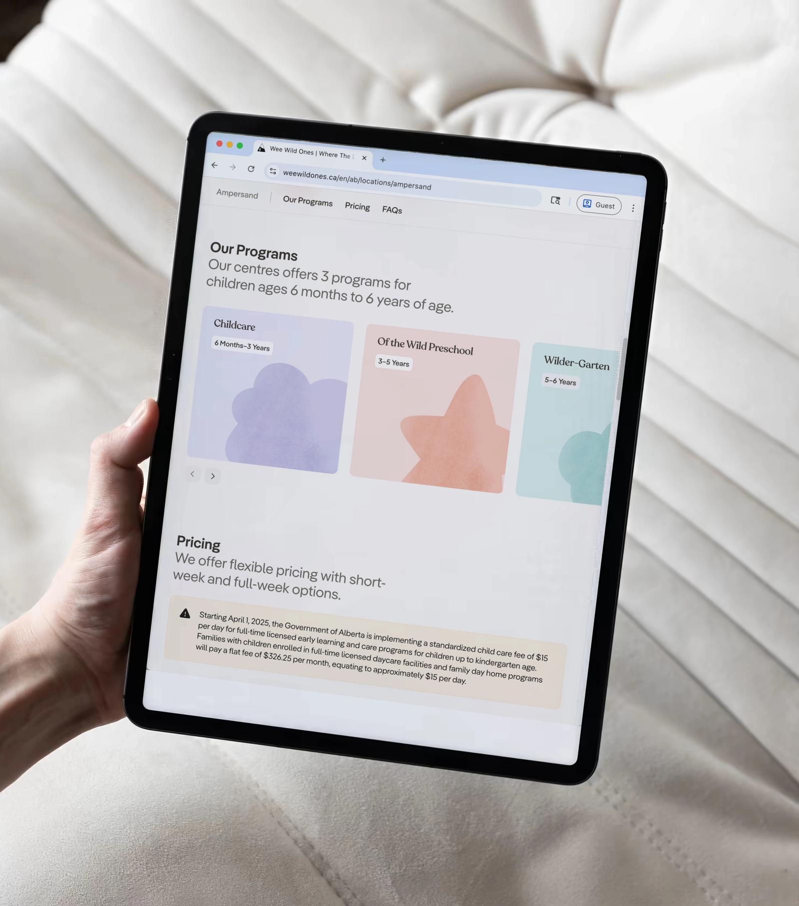 A hand holding a tablet showing the Wee Wild Ones programs and pricing sections, over a white quilted cushion.