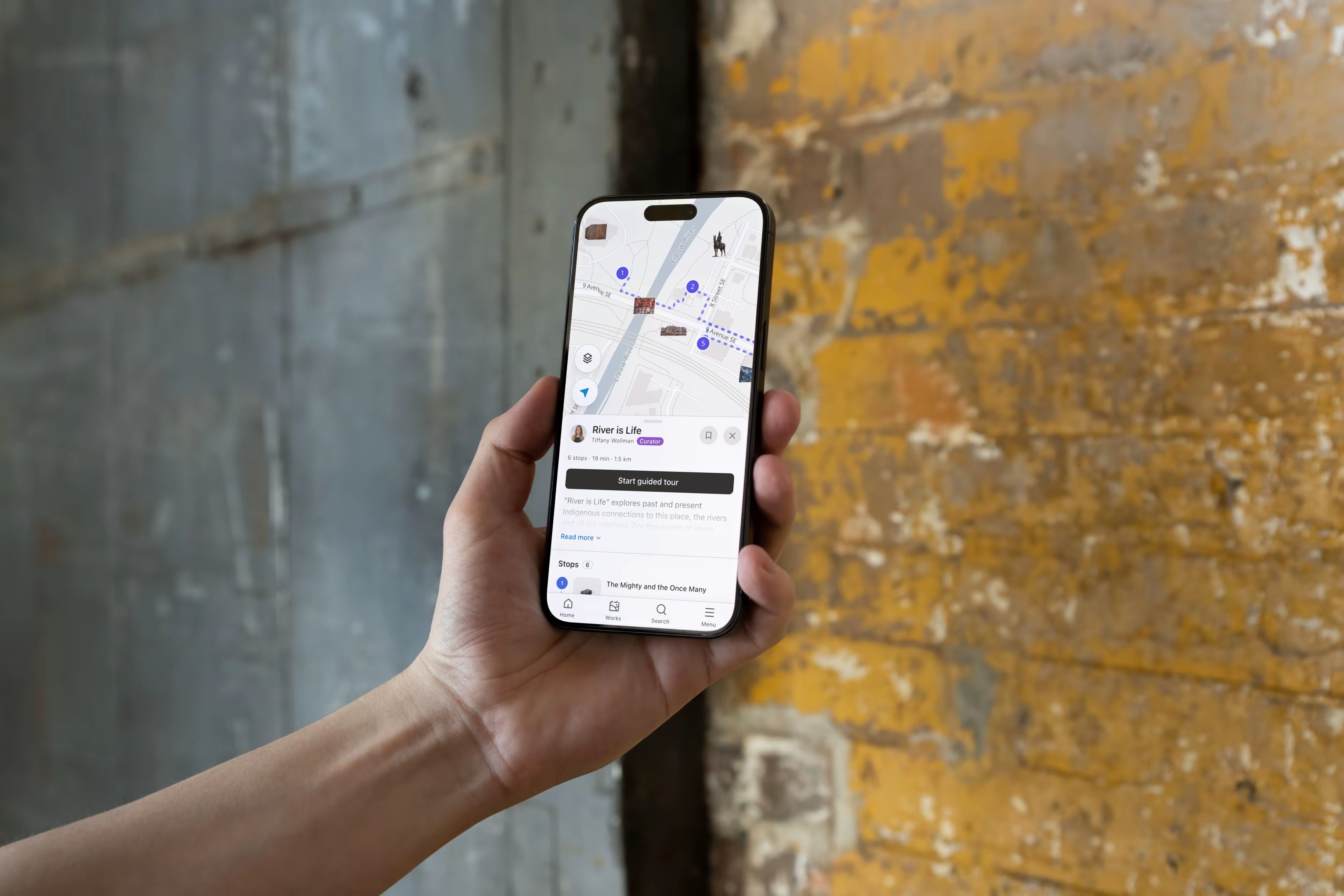 A hand holding a phone that shows the Calgary Public Art Guide map with an artwork card, against a weathered yellow and grey wall.