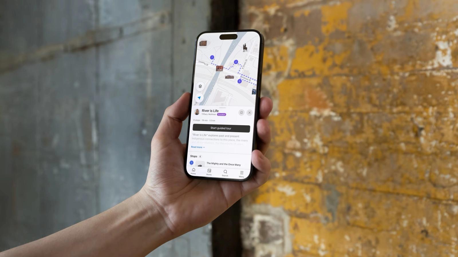A hand holding a phone that shows the Calgary Public Art Guide map with an artwork card, against a weathered yellow and grey wall.