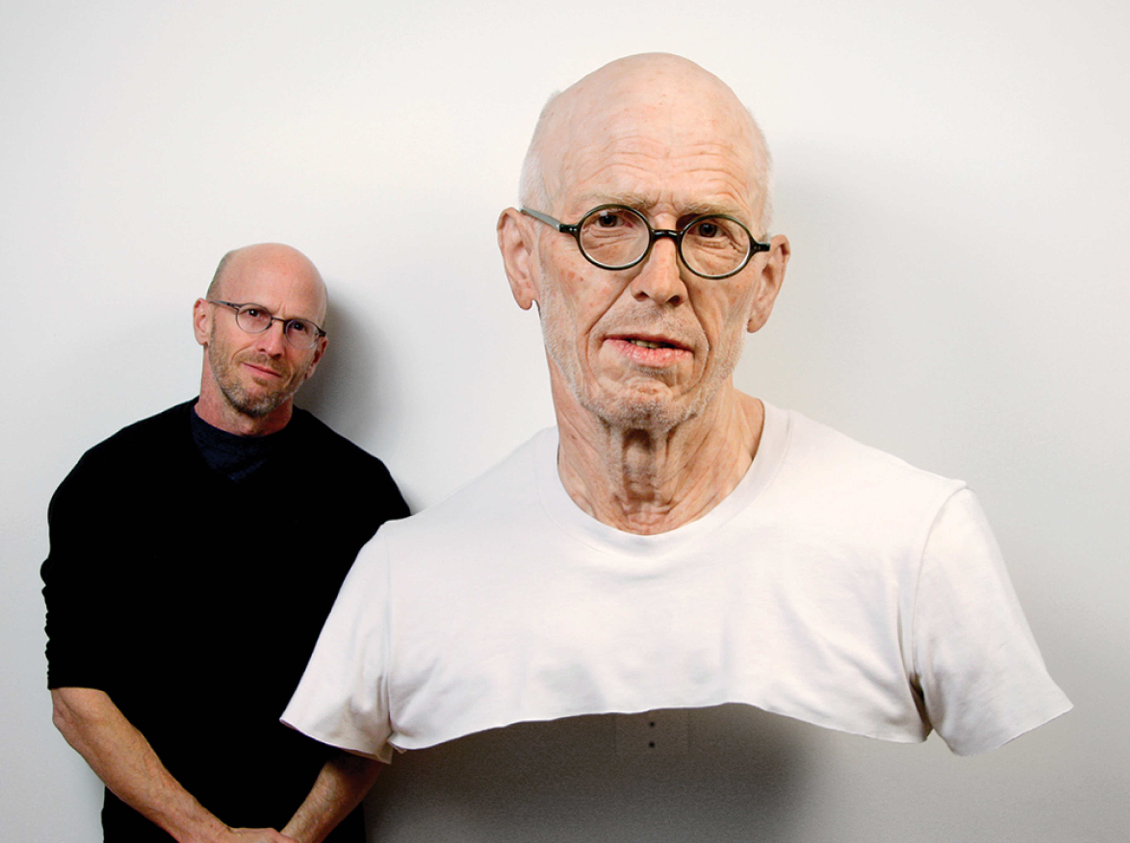 Photo of the artist standing behind a realistic sculpture of himself as an old man