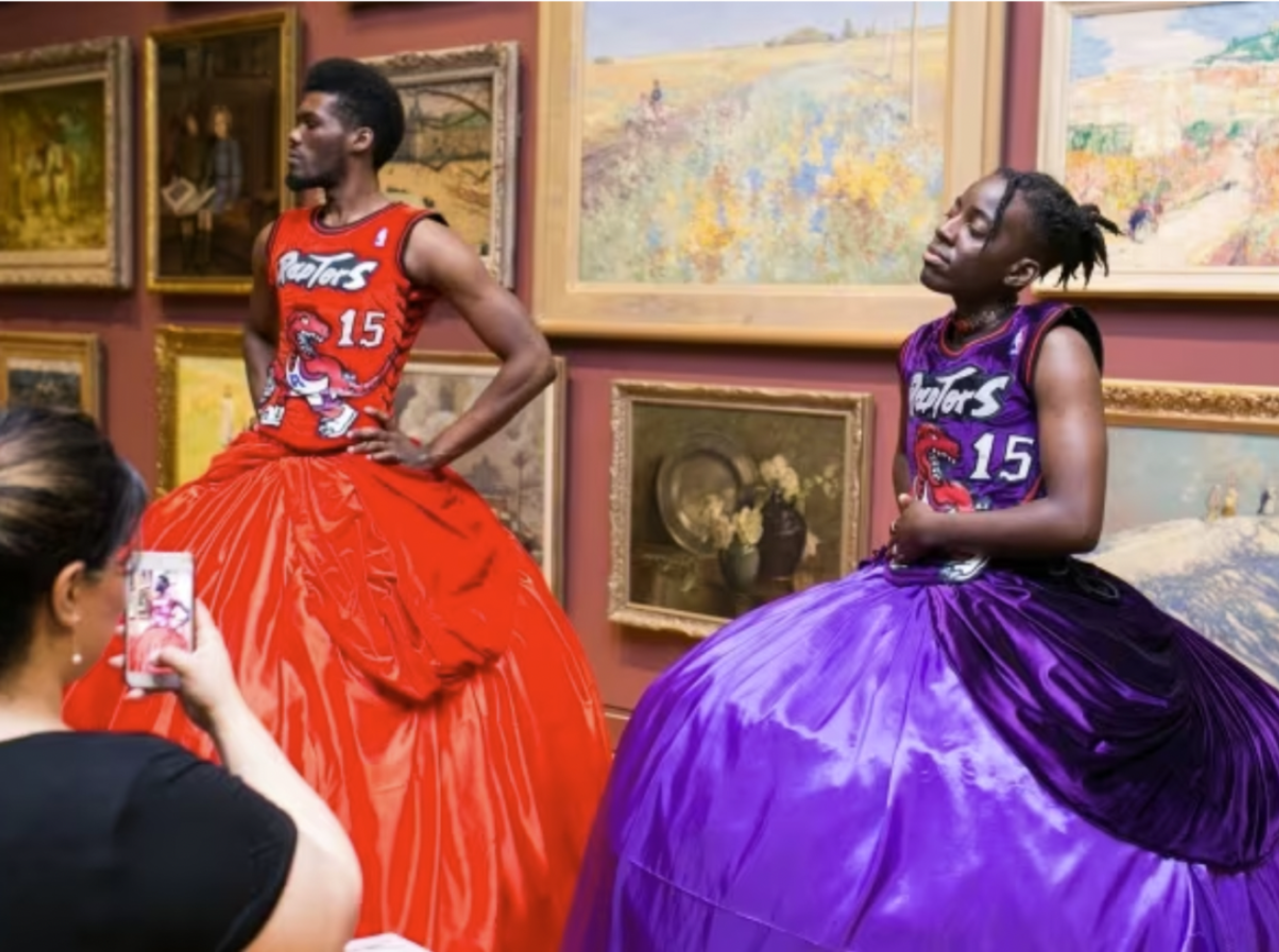 Two black models wearing one red and one purple ballgowns in front of a wall of old paintings