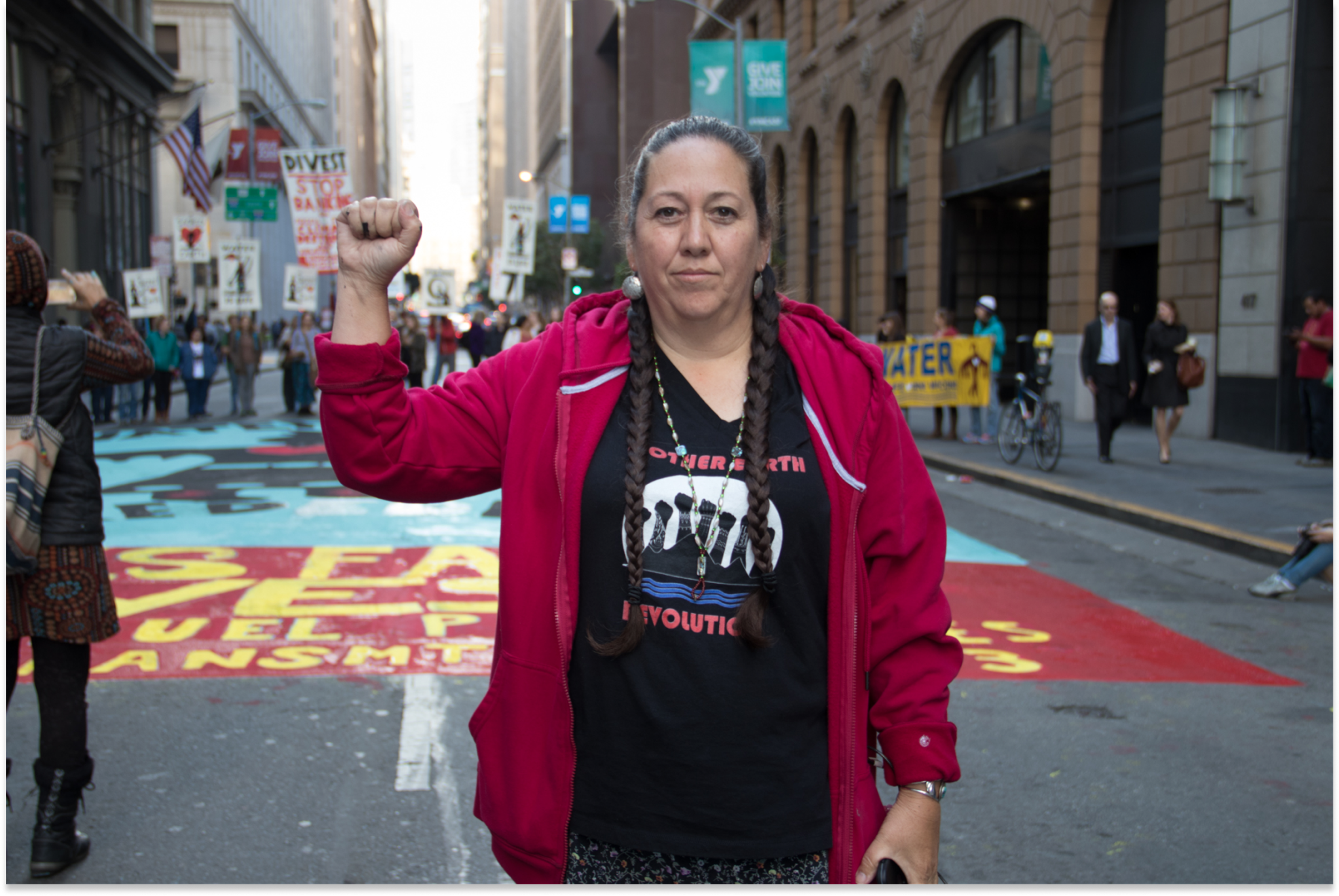 Image of Christi Belcourt in a red sweater in the street during a protest with her arm raised in a fist