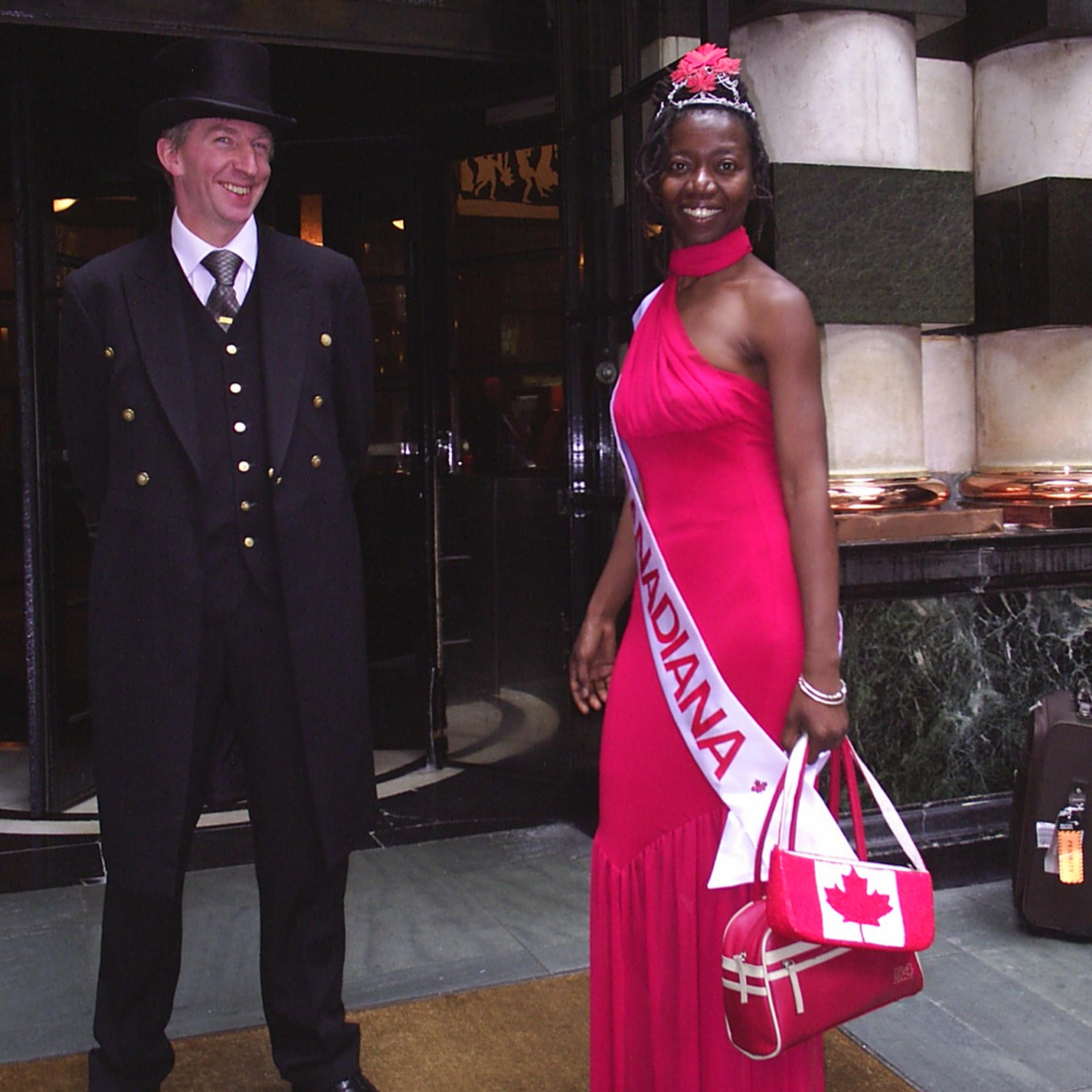 The artist Camille Turner dressed in a long red gown with a Miss Canadiana sash poses with a door man wearing a suit.