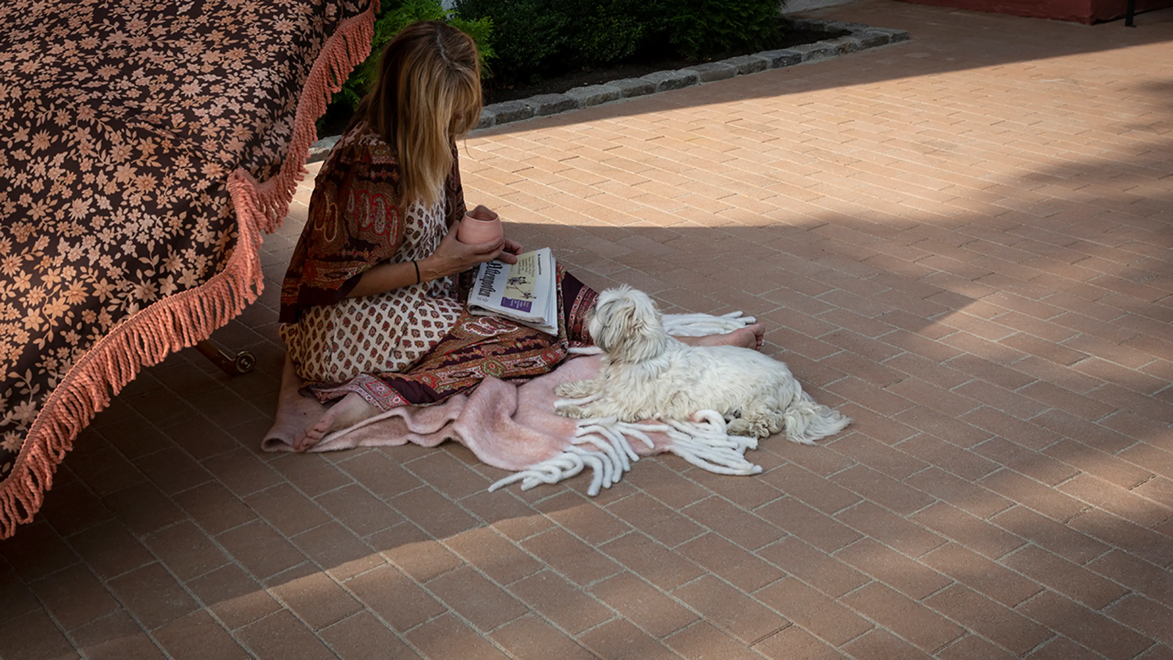 Kvinne som sitter på bakken under parasoll med en hund på uteplass med belegningsstein.