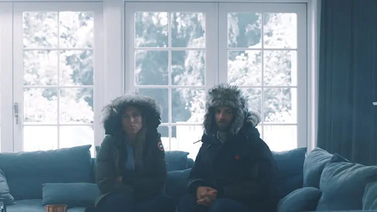 et par sitter fullt påkledd med vinterjakker innendørs i en sofa