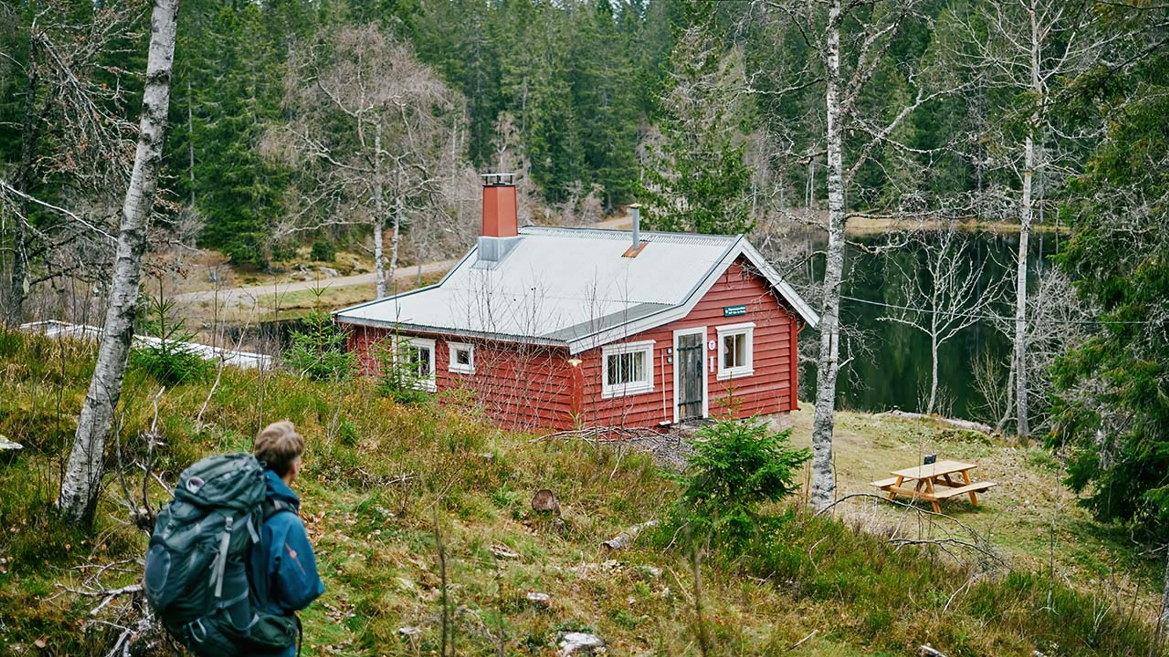 Rød hytte ved et vann i skogen og en mann med ryggsekk.