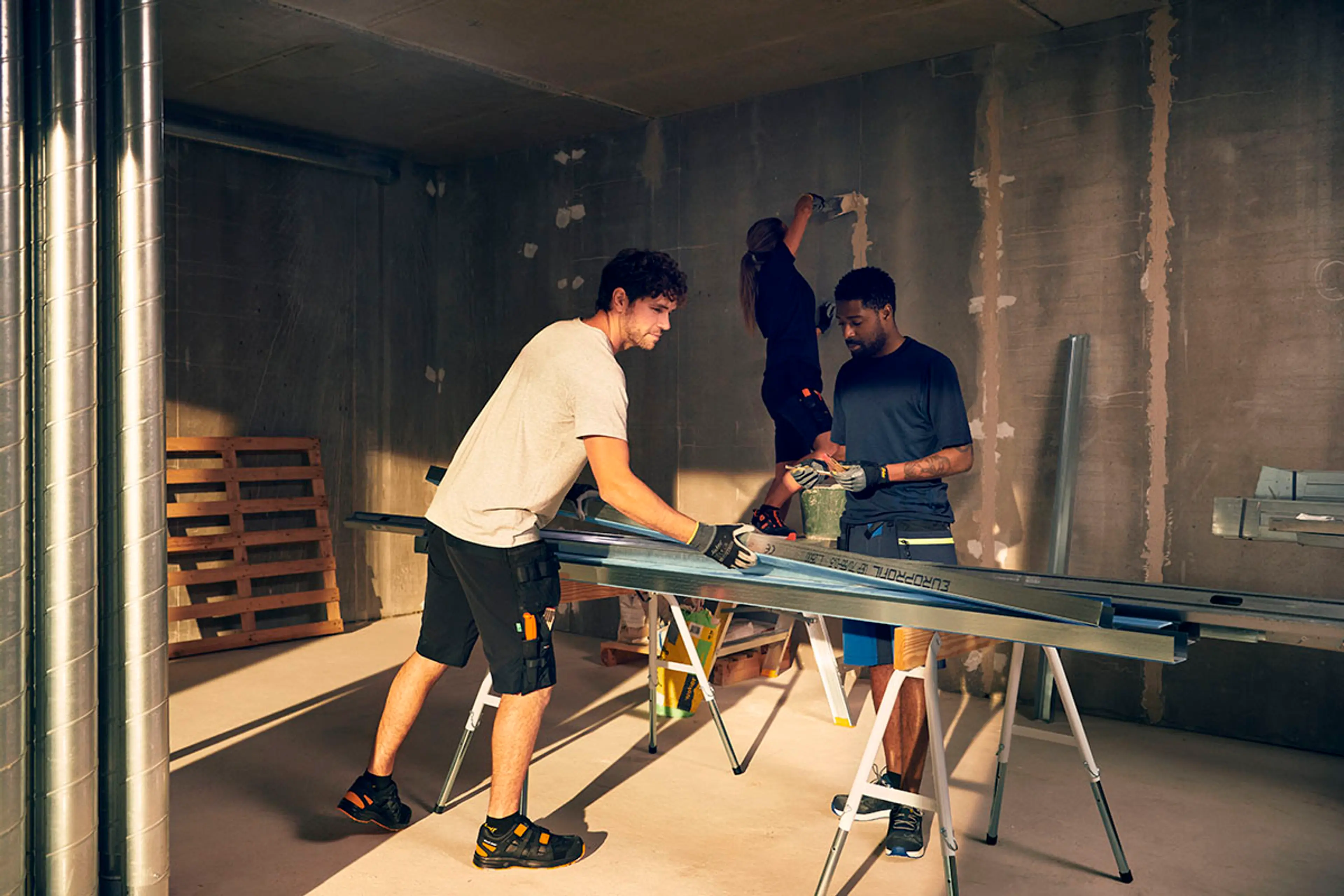 Tre håndverkere i shorts og t-skjorte jobber på en byggeplass