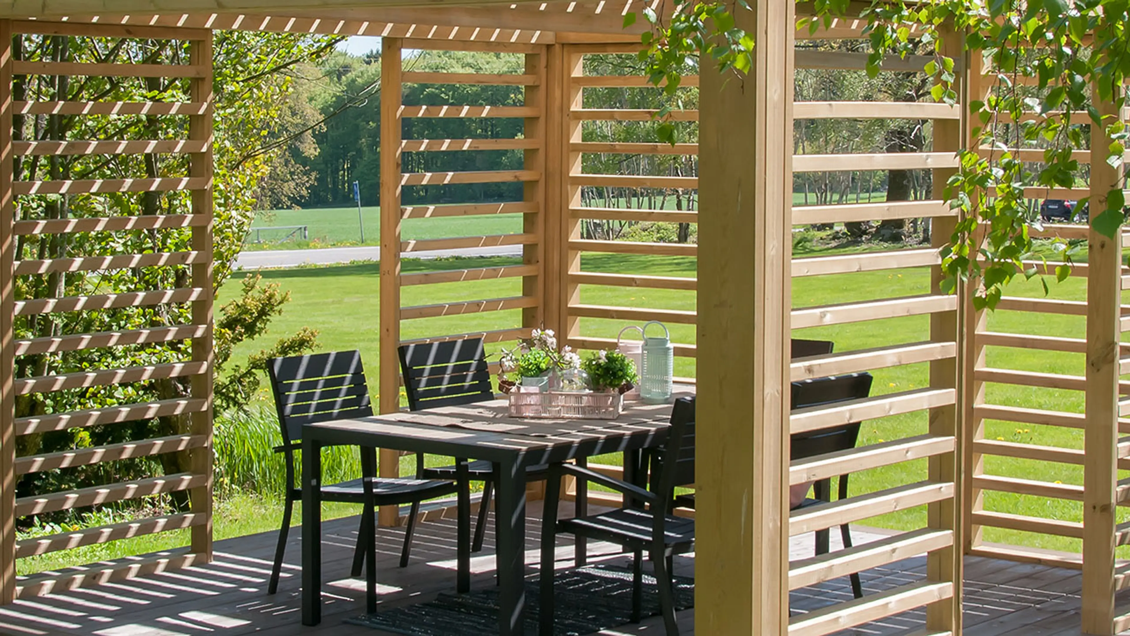 Nærbilde av stor frittstående pergola i lyst treverk med spisegruppe. 