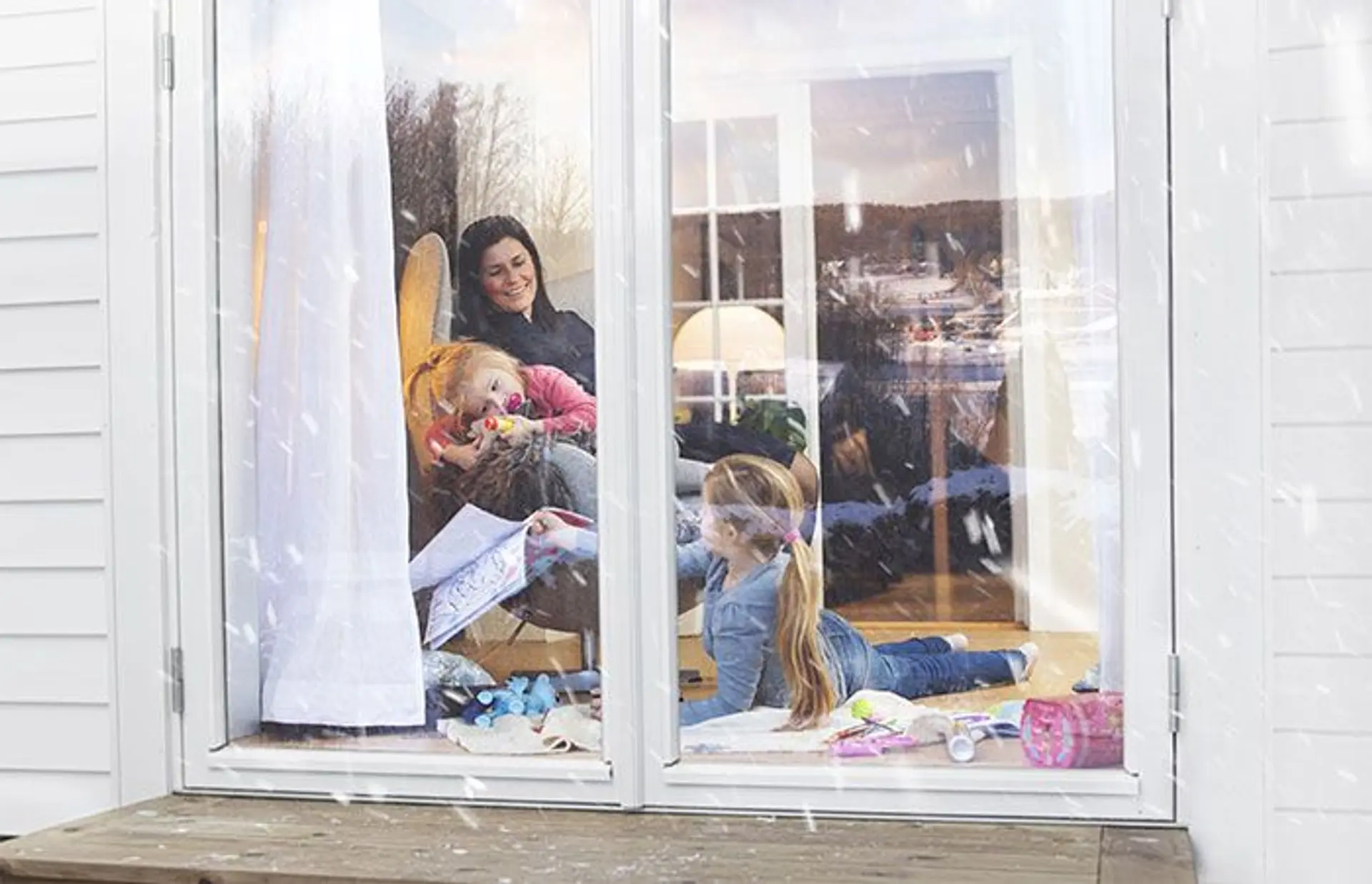 En mor og to små barn i en stue kan ses gjennom et vindu