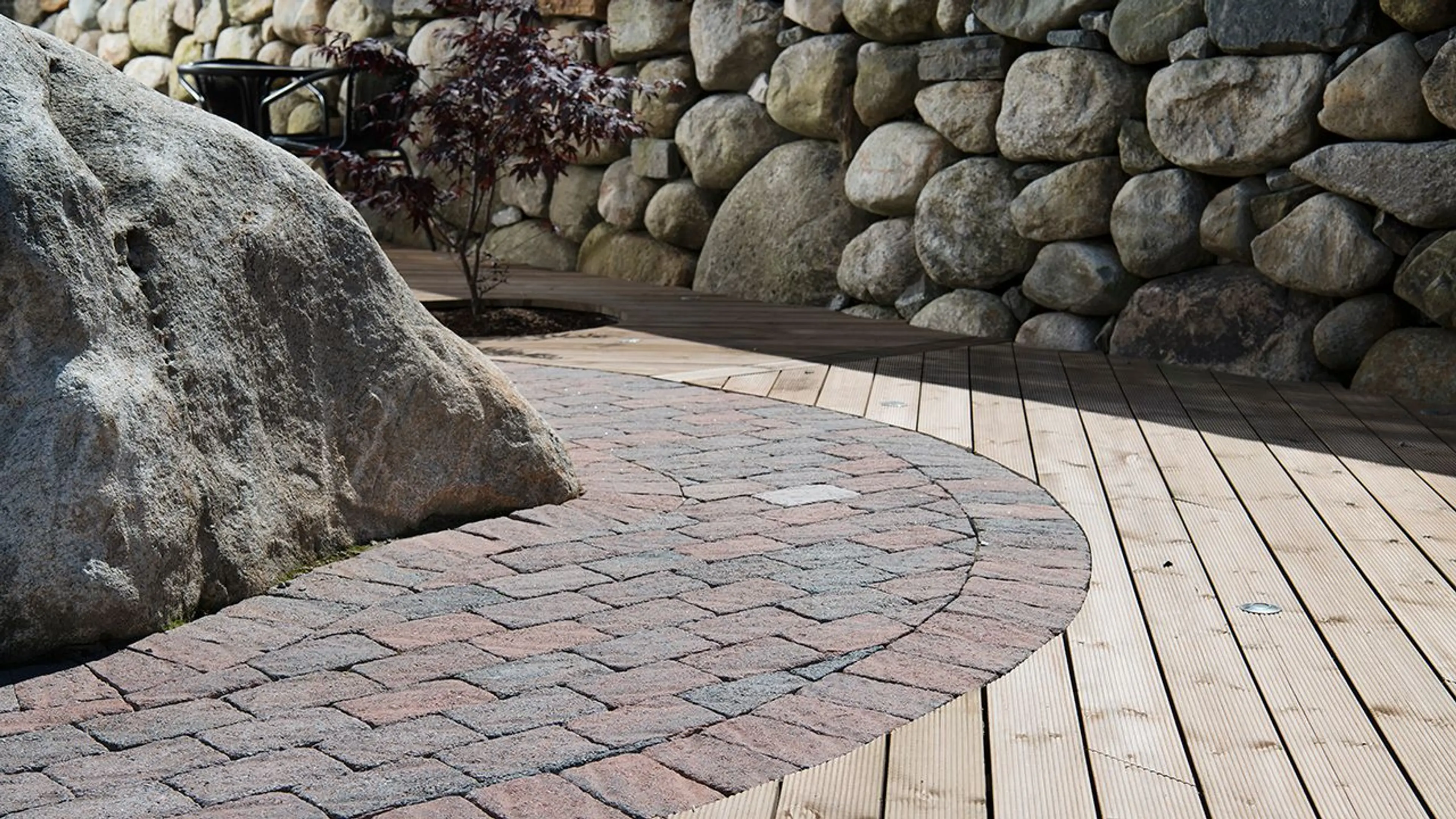 Brun belegningsstein fra Multiblokk lagt rundt naturlig stein i rund form kombinert med lyse terrassebord med innfelt utebelysning.