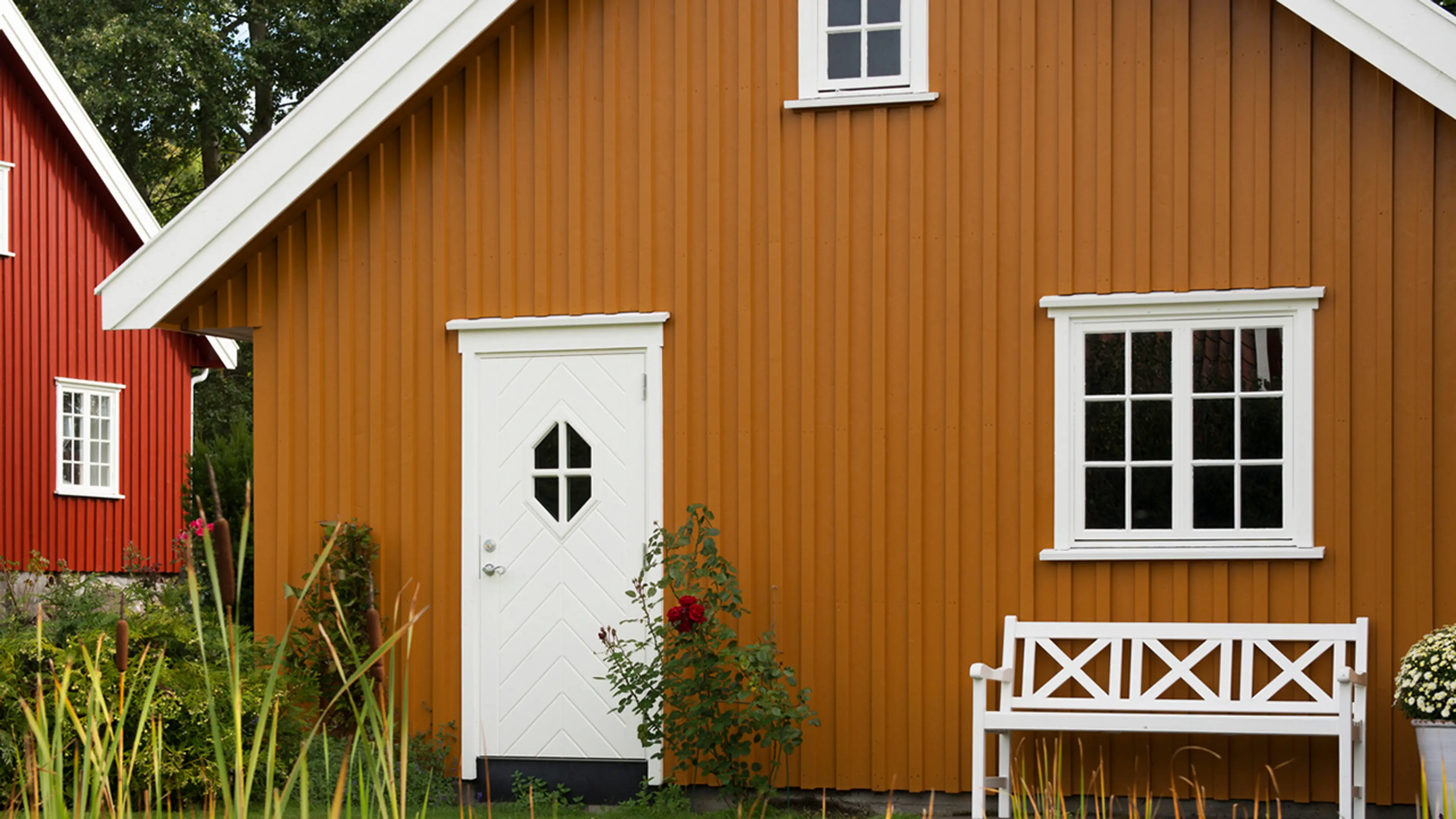 Ny kledning på gammelt hus i gult med hvite karmer og dører og rødt hus i bakgrunnen.