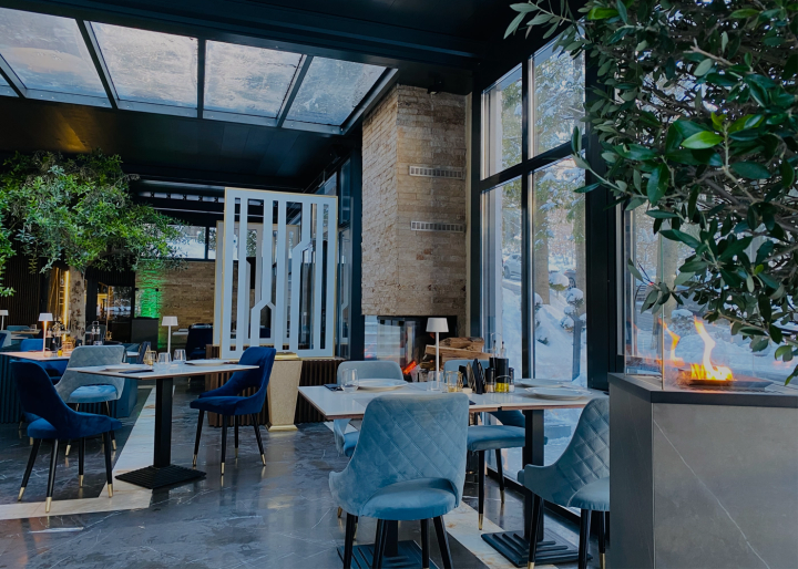 Restaurant cu acoperiș din sticlă, scaune albastre din catifea, măslin decorativ și șemineu cu bioetanol