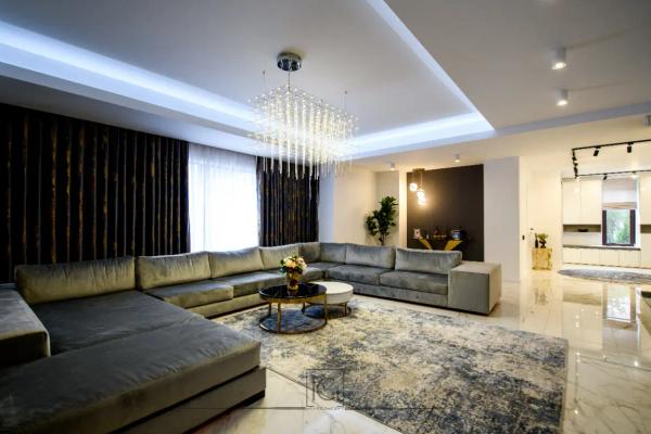 Living cu canapea gri în formă de U, covor albastru-bej și bucătărie albă în fundal – Casa M.I., Bragadiru