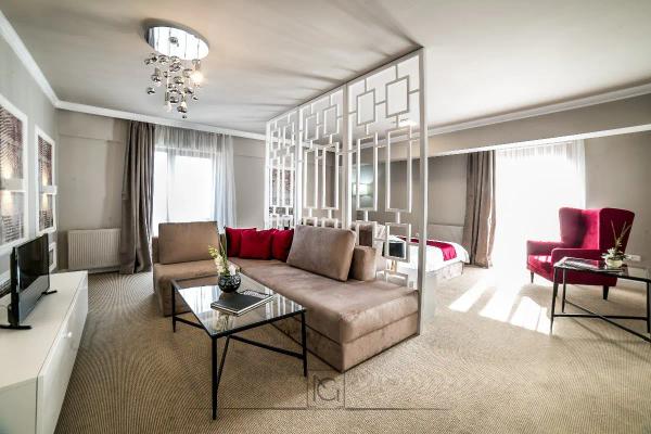 Apartament de hotel cu zonă de zi, canapea bej, fotoliu roșu și paravan geometric alb