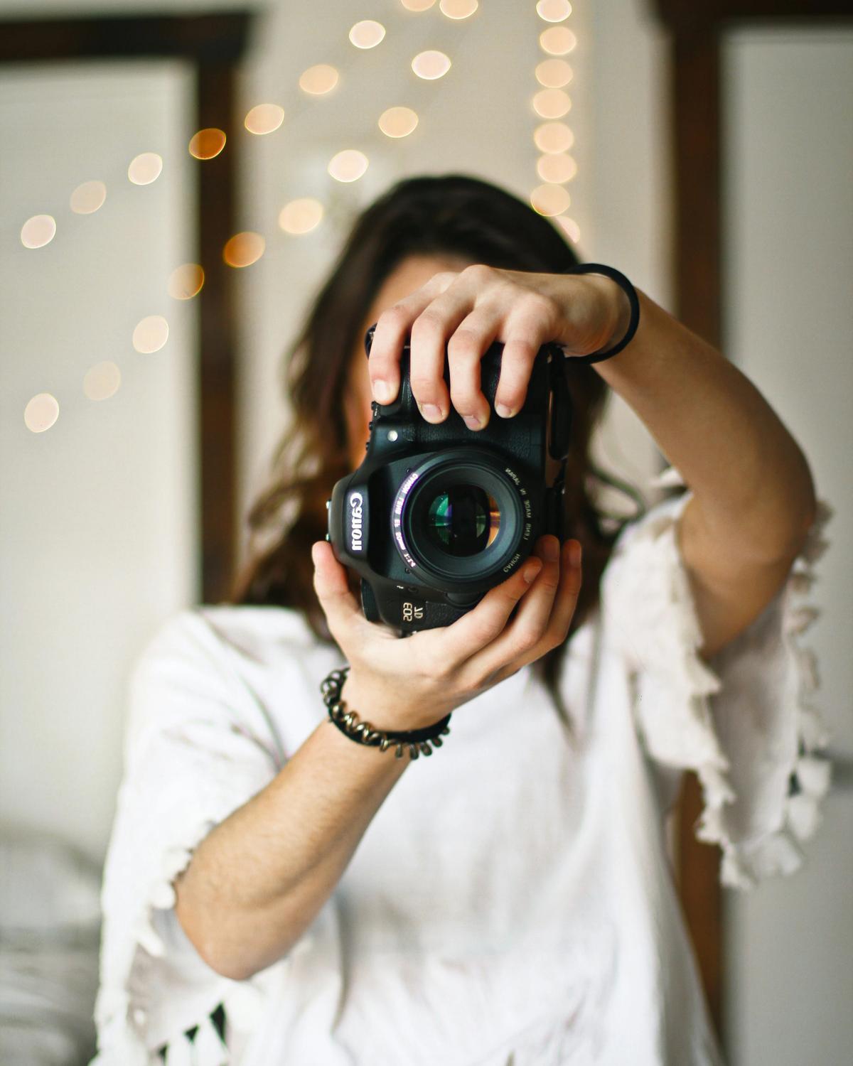 Photographer holding a camera to her eye in front of warm string lights