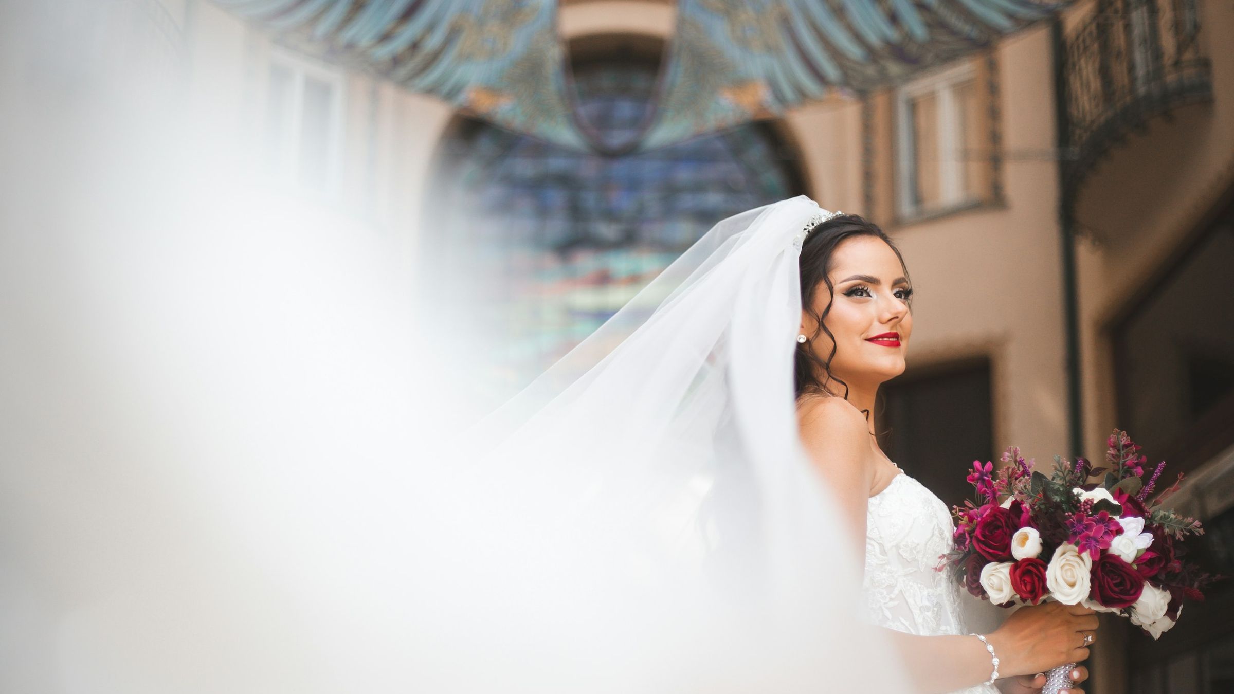 Bride with a long veil holding a red and white bouquet on a city street