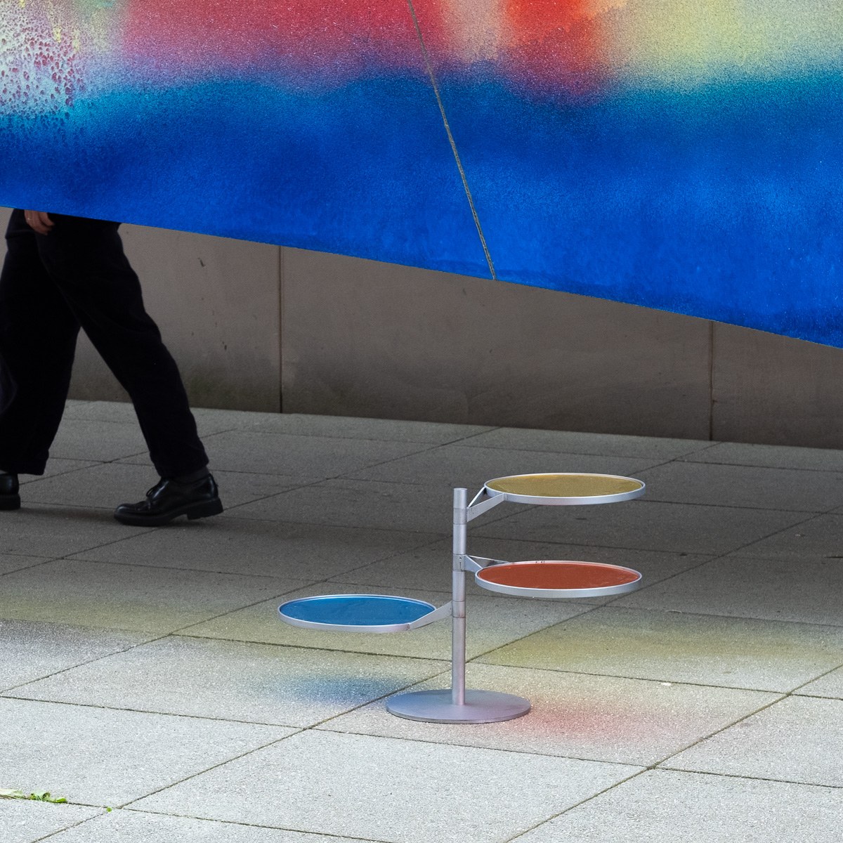 FLTR by NOEMAR — the piece on a plaza with a passer-by walking behind a large blue artwork, casting coloured light on the ground.
