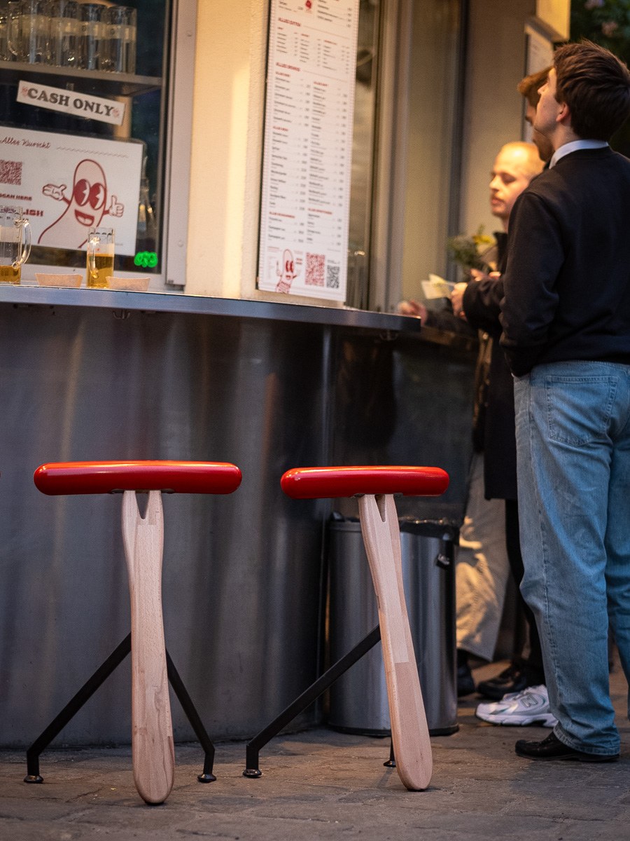Würstelhocker by NOEMAR — sausage-shaped stool balanced on a wooden fork base. Wiener Norm collection, 2026