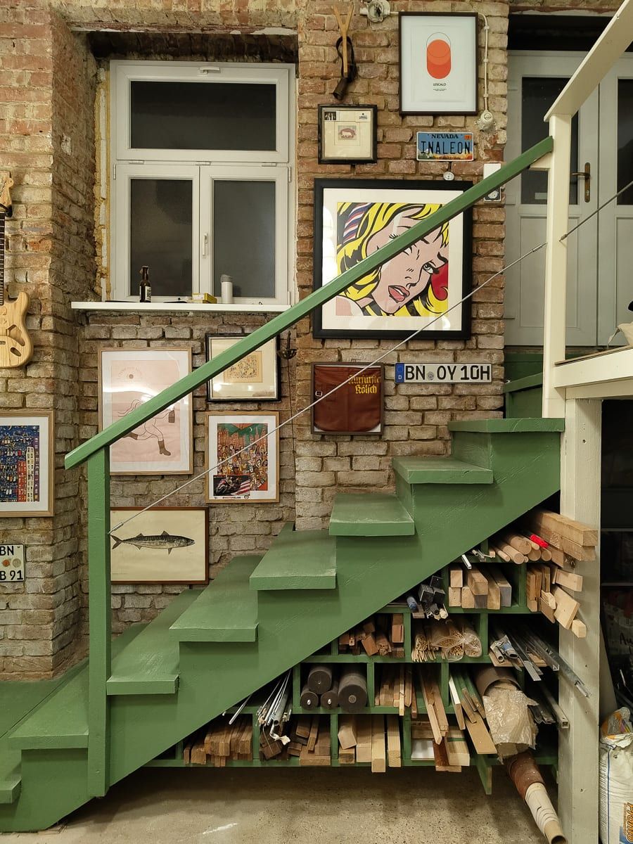 The studio's green staircase against a brick wall lined with framed prints, with timber offcuts stored in the shelving underneath.