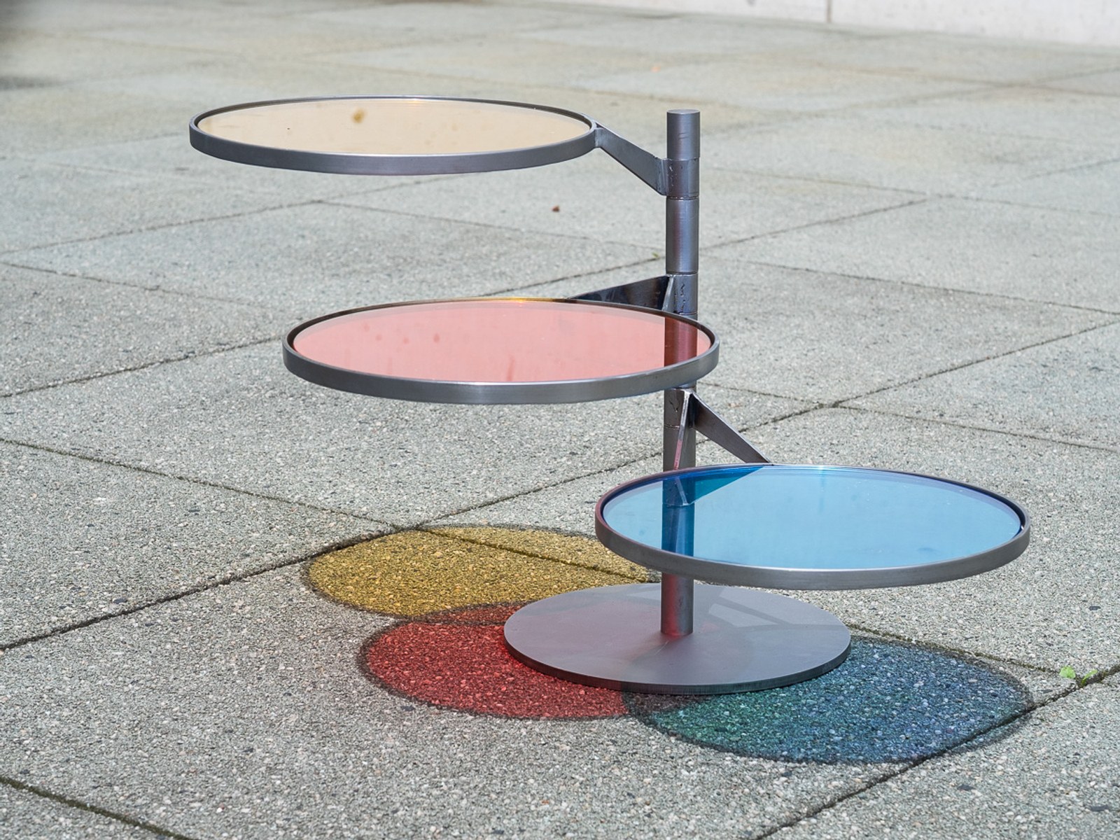 FLTR by NOEMAR — the beige, red and blue glass discs seen from above, their coloured shadows overlapping on the pavement.