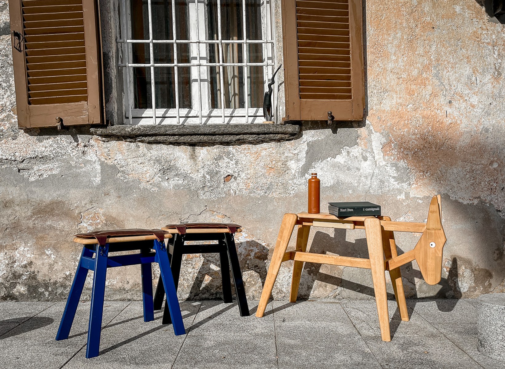 Donkey by NOEMAR — a blue and a black stool with a bare wooden frame set outside a house, styled with a bottle and books.